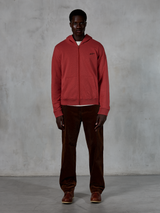 Alpinestars Essentials Ageless Zip Hoodie, casual streetwear, teglrød med lille sort stjernelogo, gennemgående lynlås foran, båret af en model med brune fløjlsbukser og støvler, eksklusivt motorsport-inspireret design
