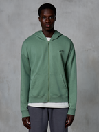 Alpinestars Essentials Ageless Zip Hoodie, afslappet streetwear-hættetrøje med lynlås, Cactus grøn, med et diskret broderet brystlogo, opdelte kængurulommer og ribbede manchetter, båret af en mandlig model mod en tekstureret grå baggrund
