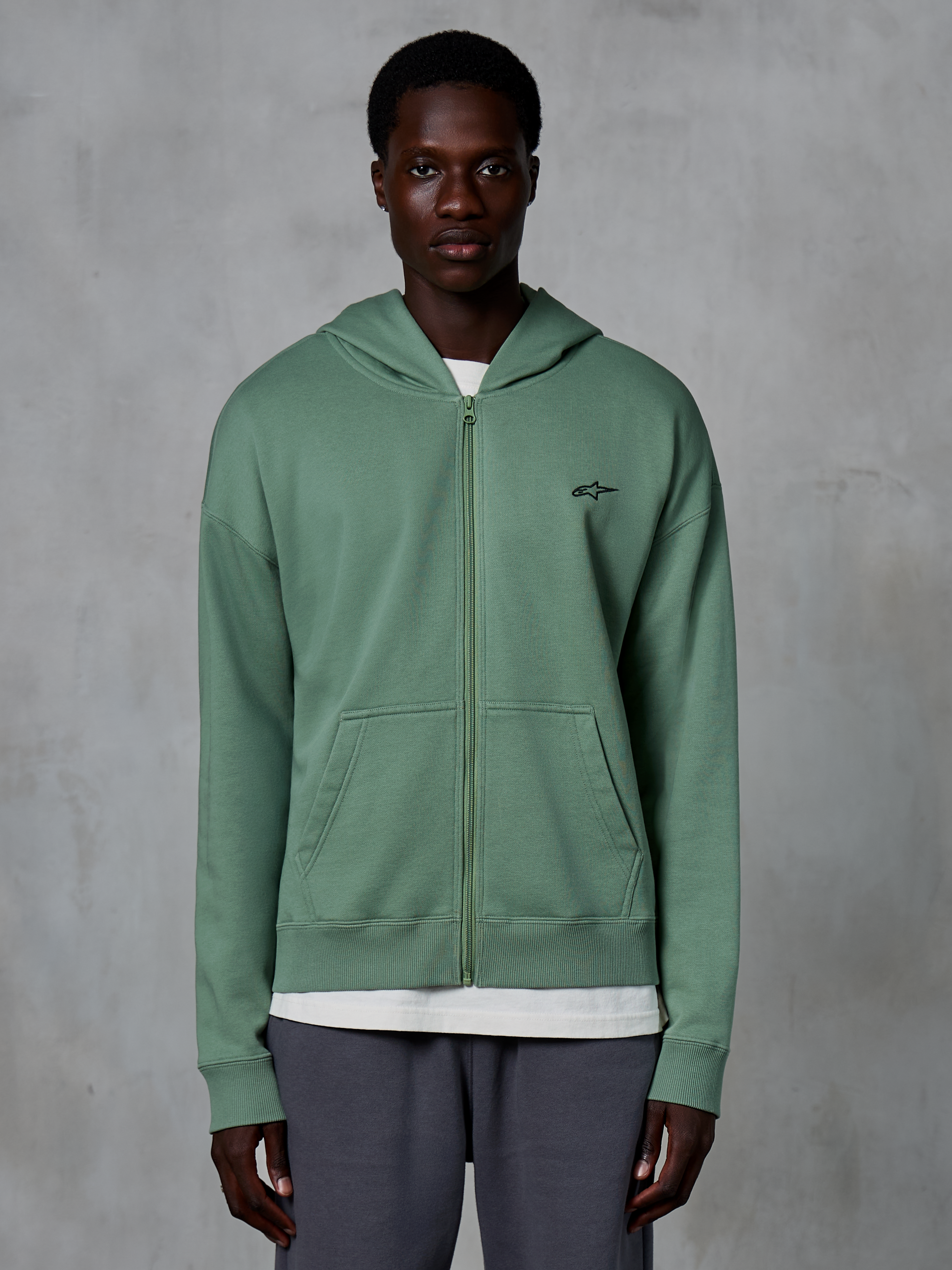 Alpinestars Essentials Ageless Zip Hoodie, afslappet streetwear-hættetrøje med lynlås, Cactus grøn, med et diskret broderet brystlogo, opdelte kængurulommer og ribbede manchetter, båret af en mandlig model mod en tekstureret grå baggrund