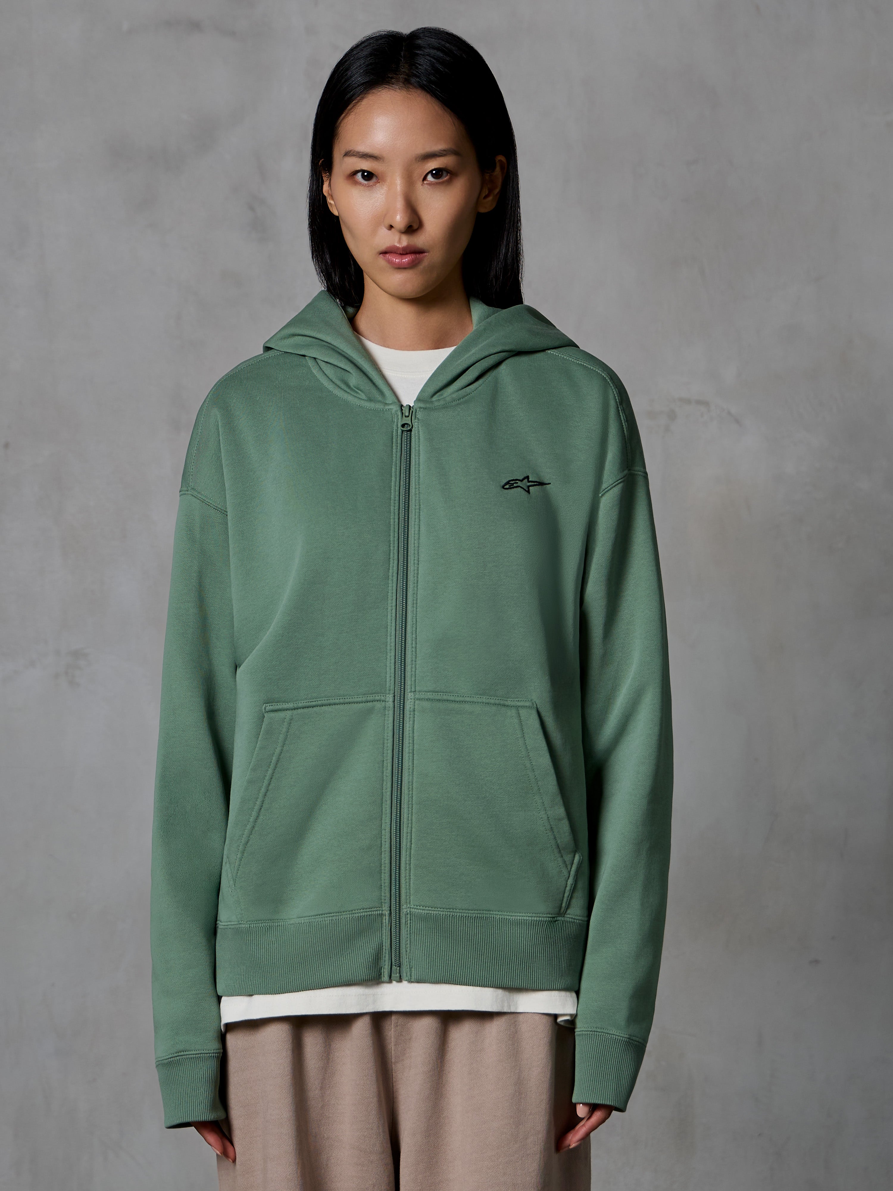 Alpinestars Essentials Ageless Zip Hoodie, kaktusgrøn, afslappet streetwear båret af en kvindelig model, fleece-hættetrøje med lynlås, lille sort logo på brystet, frontlommer, afslappet pasform til hverdagsbrug