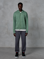 Alpinestars Essentials Ageless Zip Hoodie, hættetrøje med lynlås, kaktusgrøn, båret af en model med mørkegrå joggingbukser og sorte sneakers, med et lille sort Alpinestars-logo på brystet, afslappet pasform, casual streetwear.