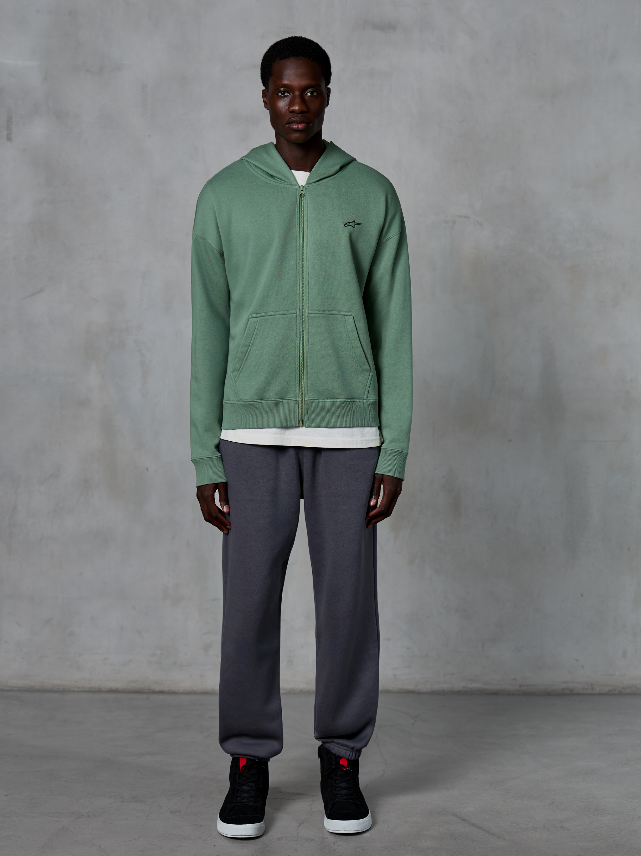 Alpinestars Essentials Ageless Zip Hoodie, hættetrøje med lynlås, kaktusgrøn, båret af en model med mørkegrå joggingbukser og sorte sneakers, med et lille sort Alpinestars-logo på brystet, afslappet pasform, casual streetwear.