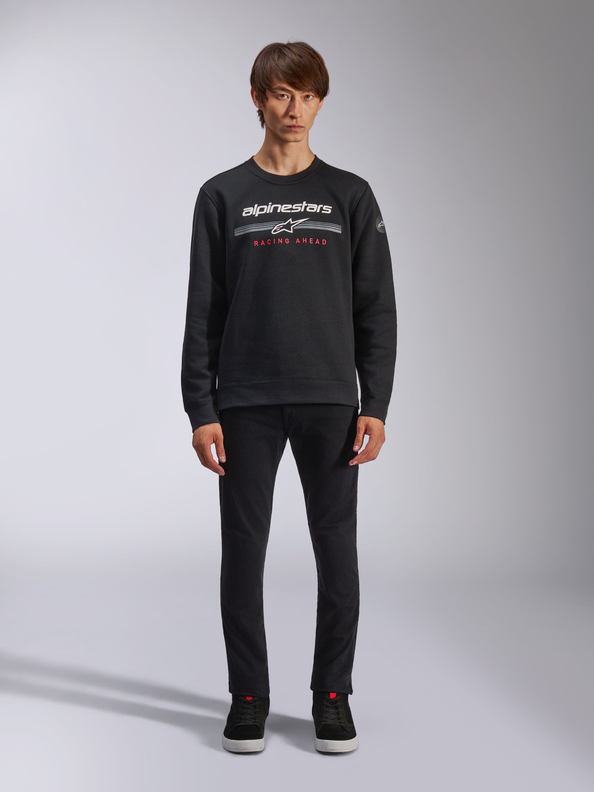 Alpinestars Bettern Crew, Black Crew Neck Sweatshirt, features white alpinestars logo with grey horizontal stripes and red 'RACING AHEAD' text, casual motorsport streetwear, worn by model with black pants and sneakers