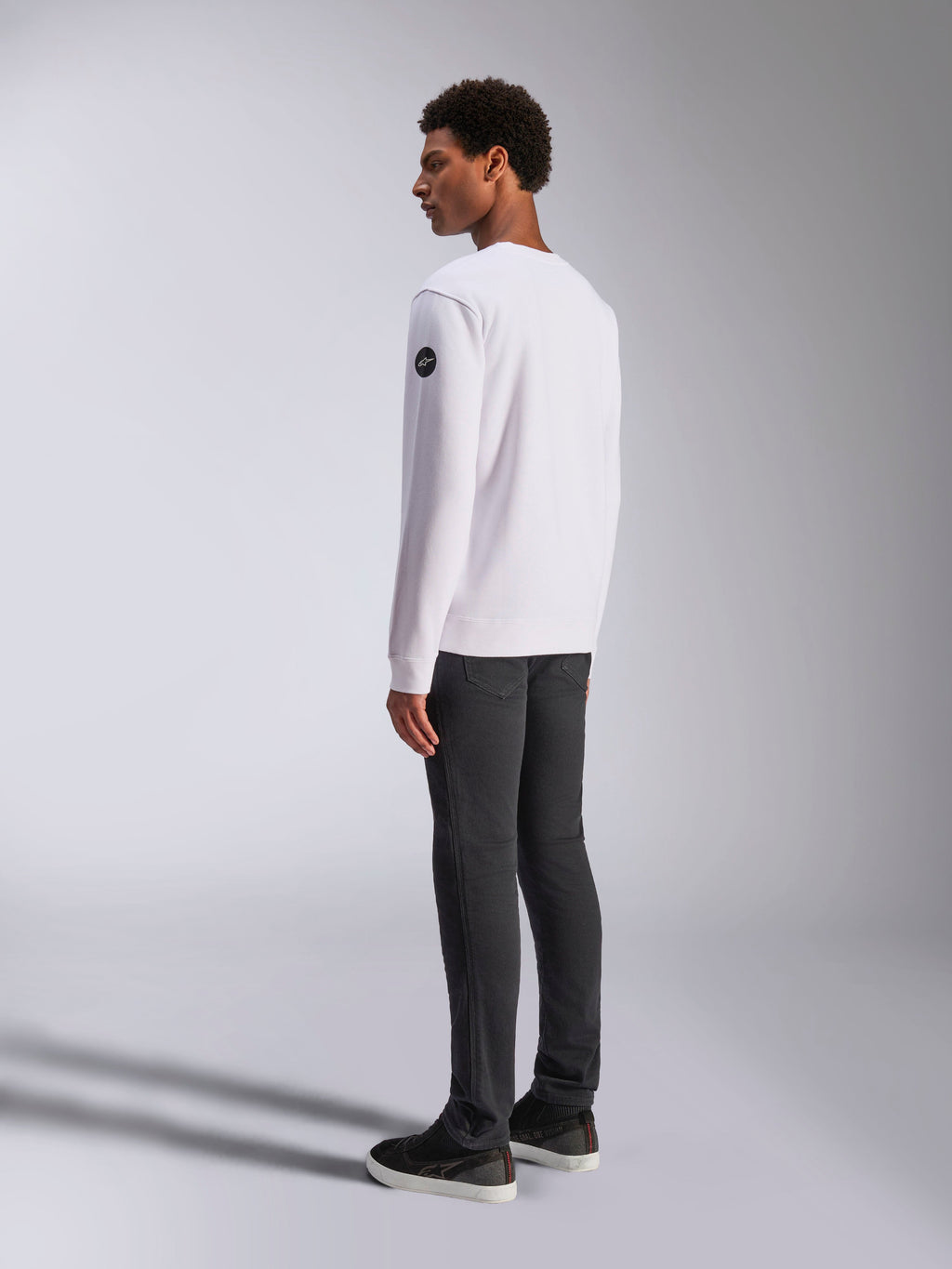 Alpinestars Bettern Crew, White Crewneck Sweatshirt, back and side view, black circular logo on the sleeve, worn with dark grey pants and black sneakers, casual motorsport lifestyle streetwear