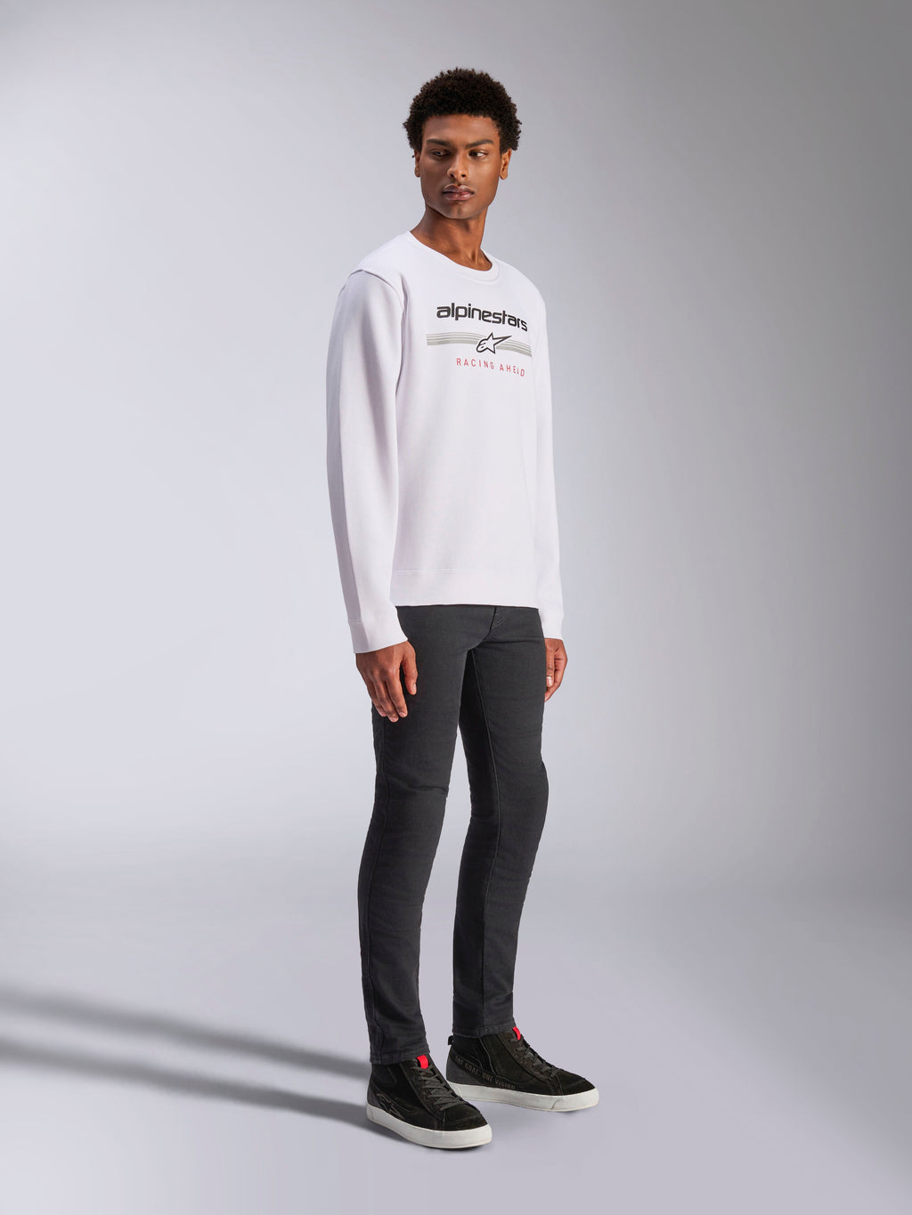 Alpinestars Bettern Crew, White Pullover Sweatshirt, featuring black Alpinestars logo and 'Racing Ahead' red text, casual motorsport streetwear, worn by a model with dark pants and black sneakers