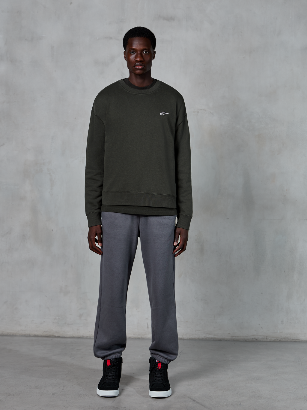Alpinestars Essentials Ageless Crew, mørkegrøn crewneck sweatshirt med et lille hvidt logo på brystet, båret af en model med grå joggingbukser og sorte high-top sneakers, afslappet livsstilstøj.