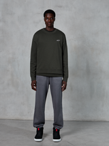Alpinestars Essentials Ageless Crew, mørkegrøn crewneck sweatshirt med et lille hvidt logo på brystet, båret af en model med grå joggingbukser og sorte high-top sneakers, afslappet livsstilstøj.