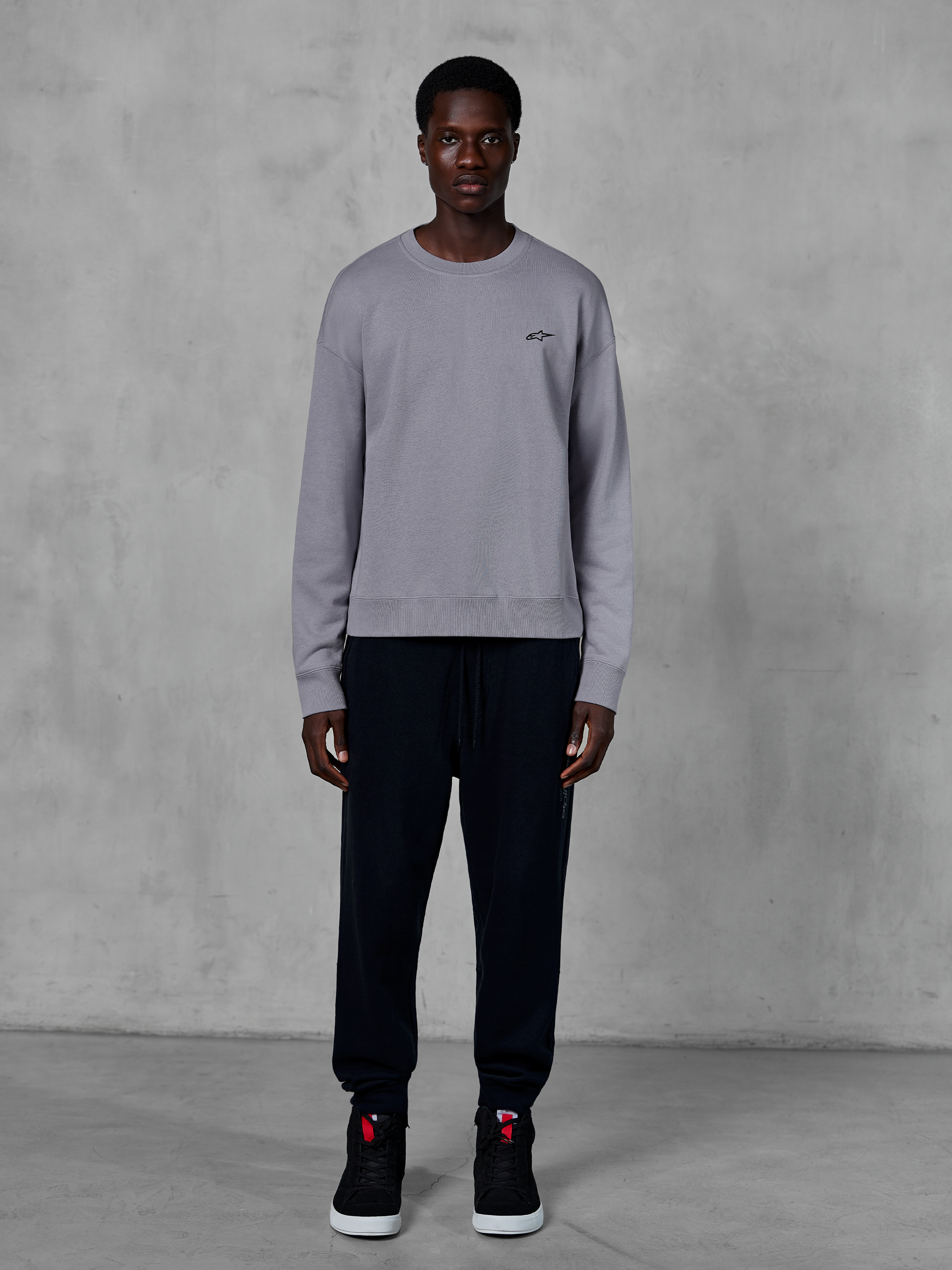 Alpinestars Essentials Ageless Crew, sølvfarvet sweatshirt med rund hals, båret af en model med sorte joggingbukser og sorte sneakers, med et lille sort Alpinestars-logo på brystet, afslappet motorsport-inspireret streetwear