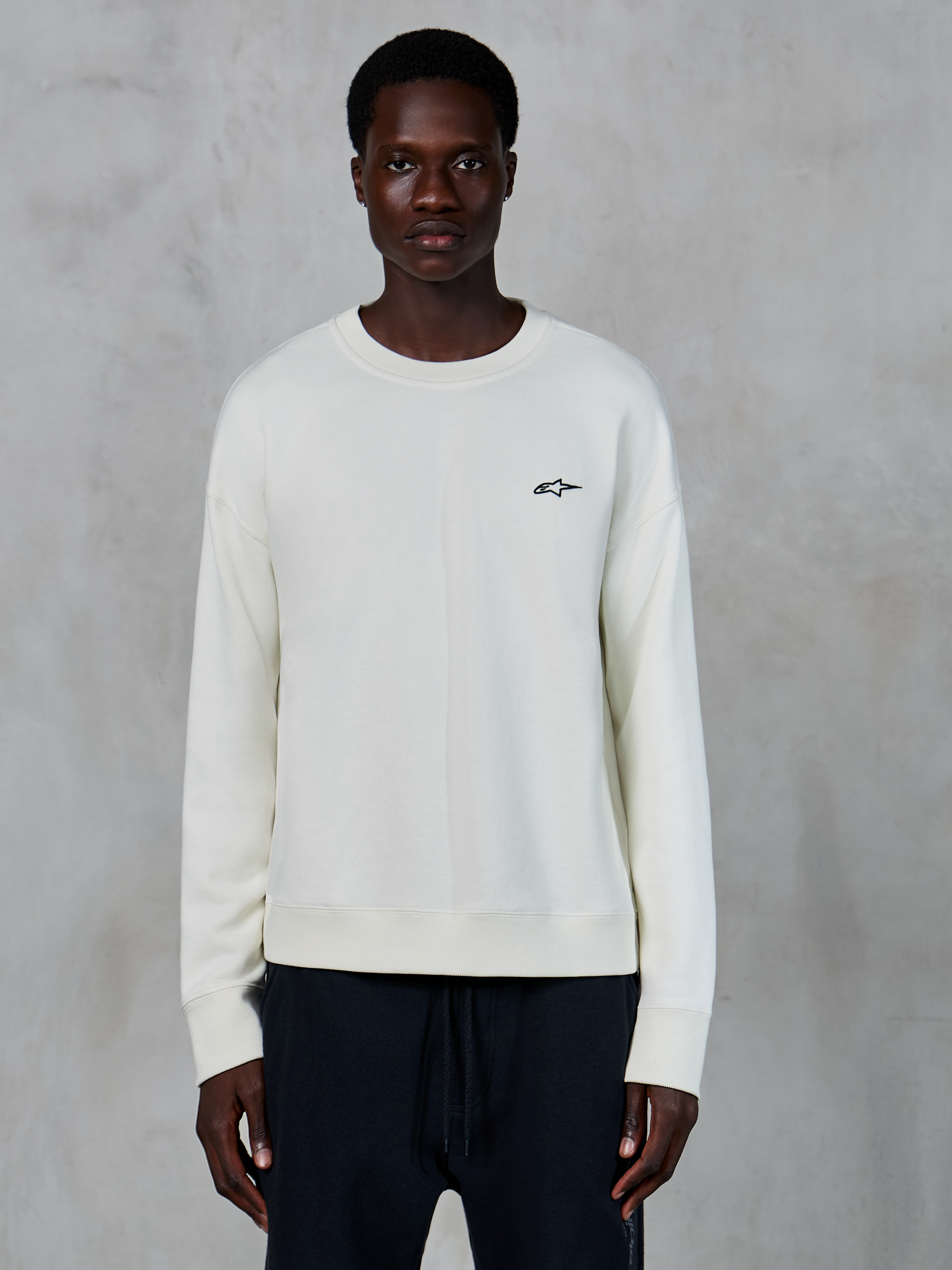 Alpinestars Essentials Ageless Crew, off-white crewneck sweatshirt, lille sort Astars-logo på venstre bryst, afslappet livsstilssportstøj, premium fleece-tekstur, båret af model med sorte joggingbukser