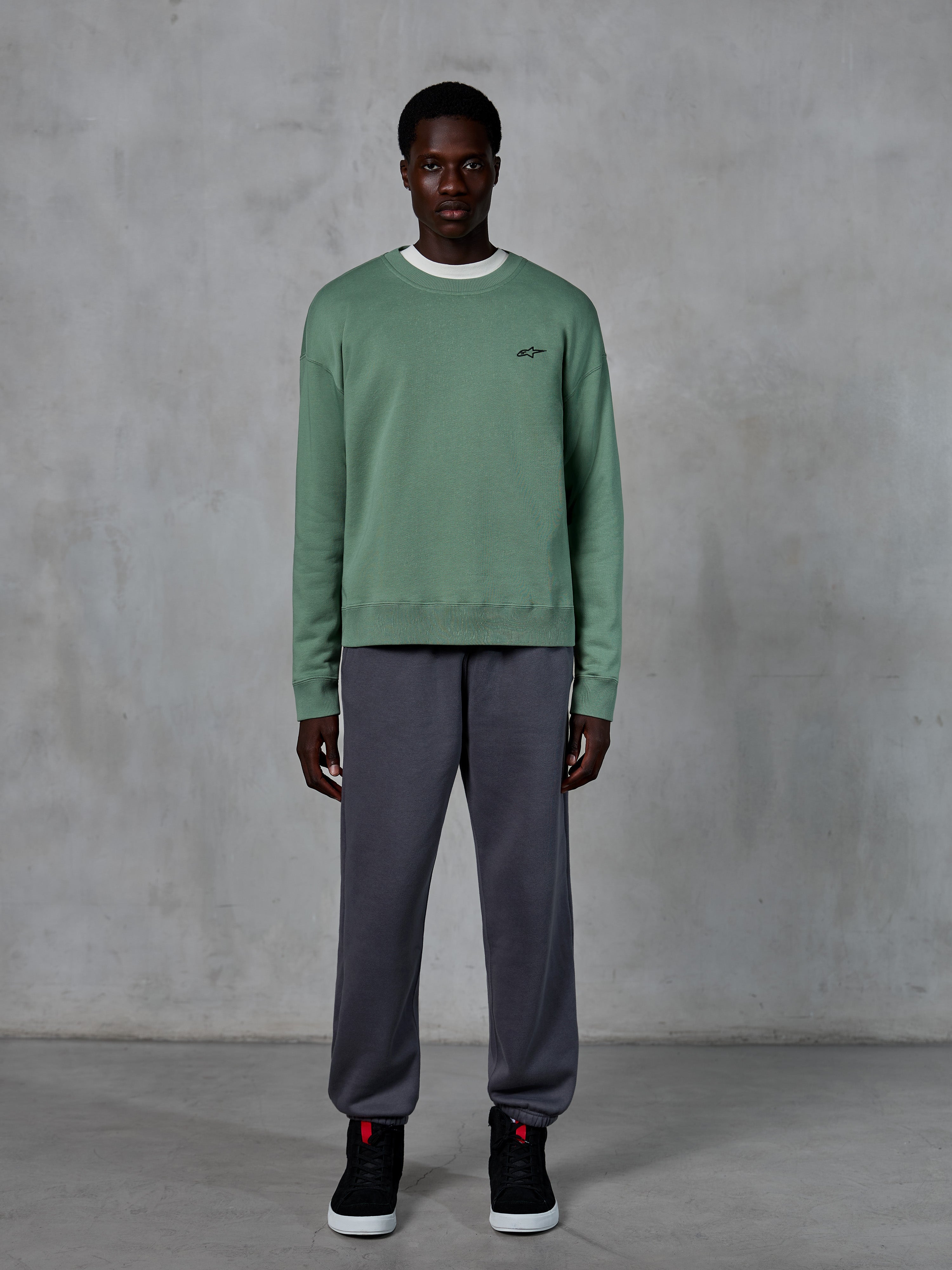 Alpinestars Essentials Ageless Crew, crewneck sweatshirt i Cactus grøn, med et lille sort brystlogo, båret af en model med grå joggingbukser og sorte sneakers, afslappet motorsport-inspireret streetwear
