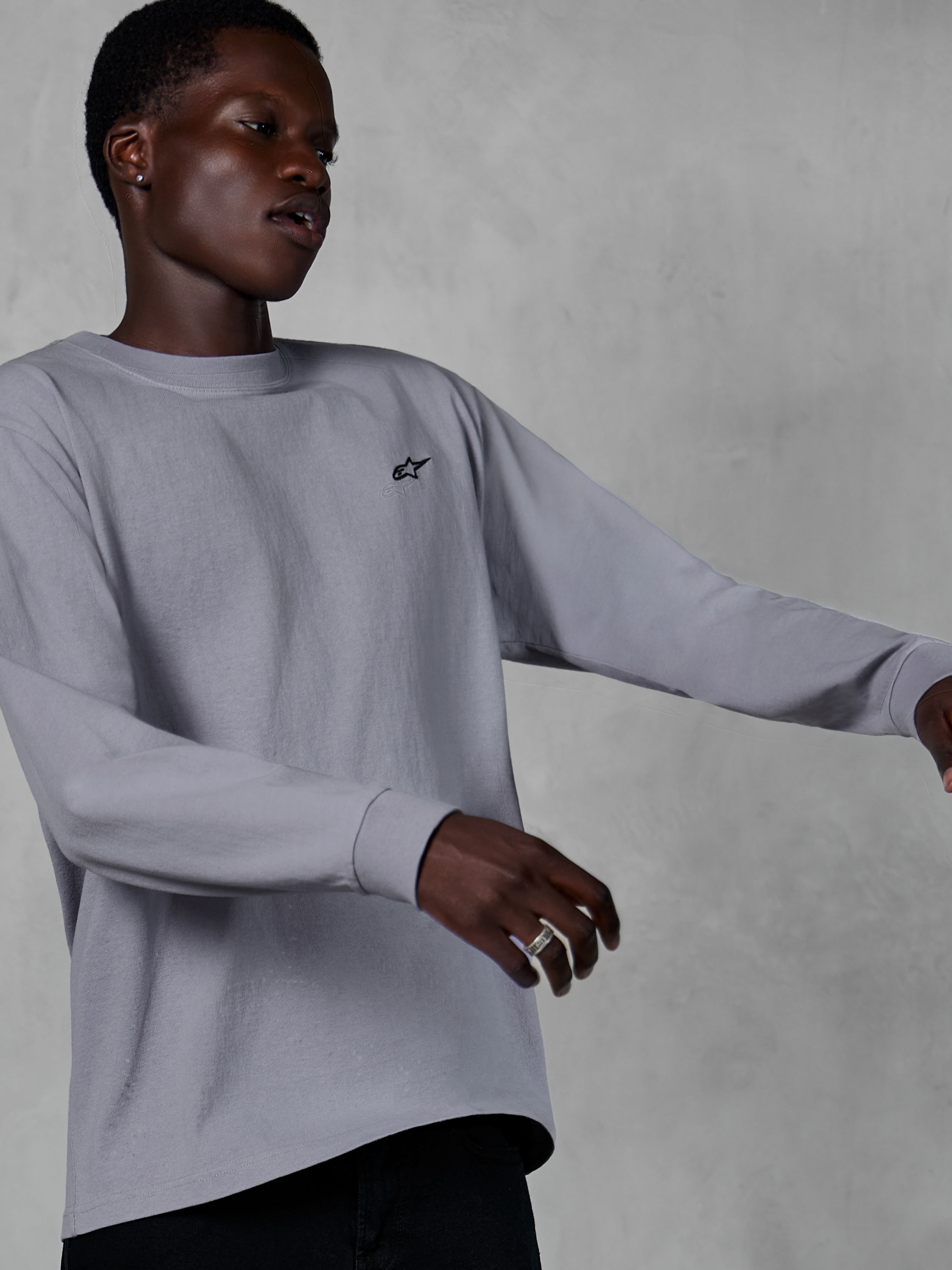 Alpinestars Essentials Ageless LS Tee, langærmet T-shirt, sølv, lille sort logo på brystet, afslappet motorsport-inspireret streetwear, båret af en model mod en grå tekstureret baggrund