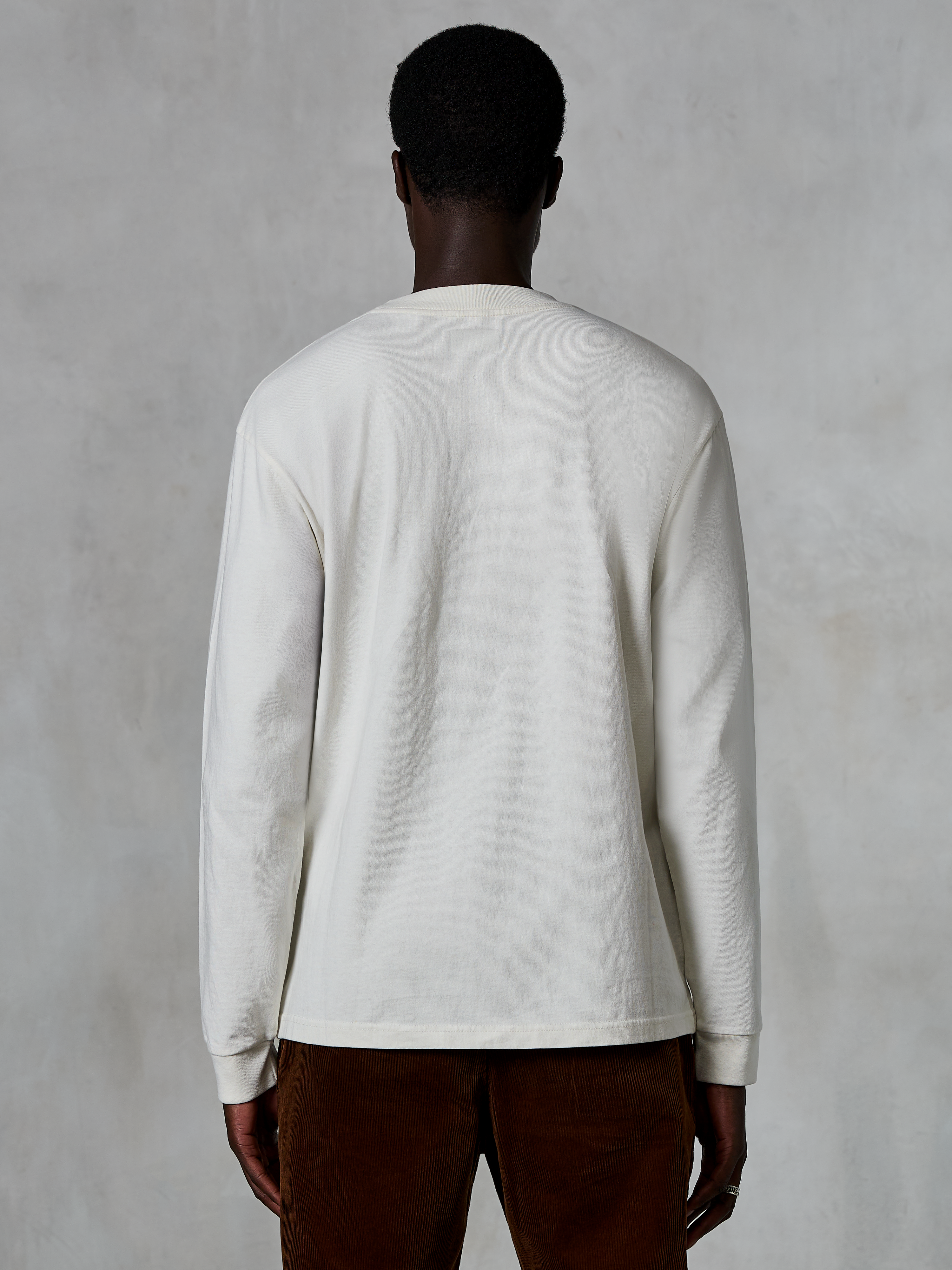 Alpinestars Essentials Ageless LS Tee, langærmet T-shirt, Off White, set bagfra på en person iført trøjen med brune fløjlsbukser, afslappet pasform, tekstureret økologisk bomuldsstof, minimalistisk afslappet streetwear