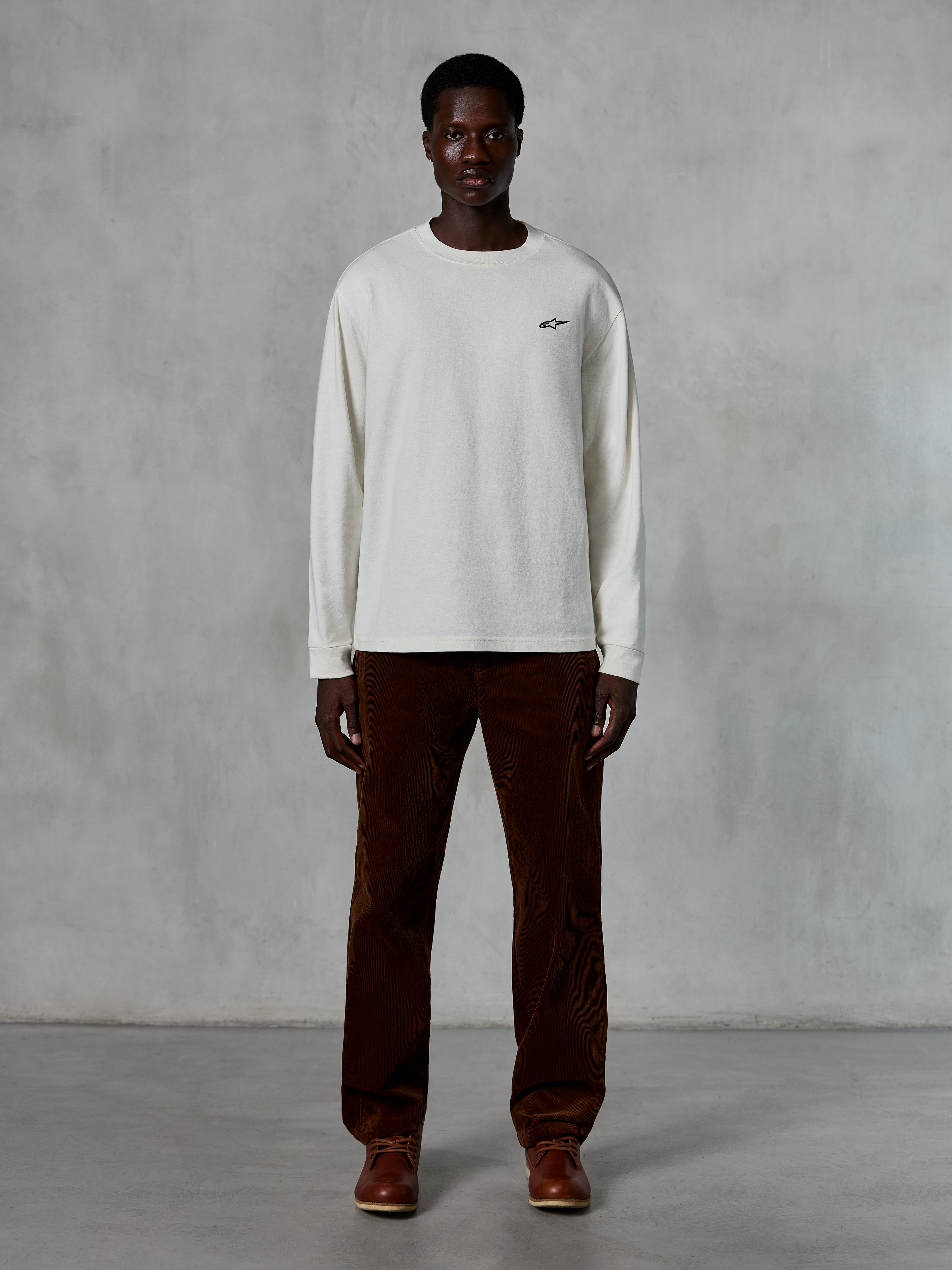 Alpinestars Essentials Ageless LS Tee, langærmet t-shirt, råhvid, båret af model med brune fløjlsbukser og brune læderstøvler, med et lille sort logo på brystet, afslappet moderne pasform, afslappet lifestyle streetwear