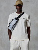 Alpinestars Essentials Ageless SS Tee, kortærmet T-shirt, råhvid, båret med matchende shorts og en sandfarvet kasket, med et lille sort logo på brystet og en grå crossbody-taske, afslappet motorsport-inspireret streetwear