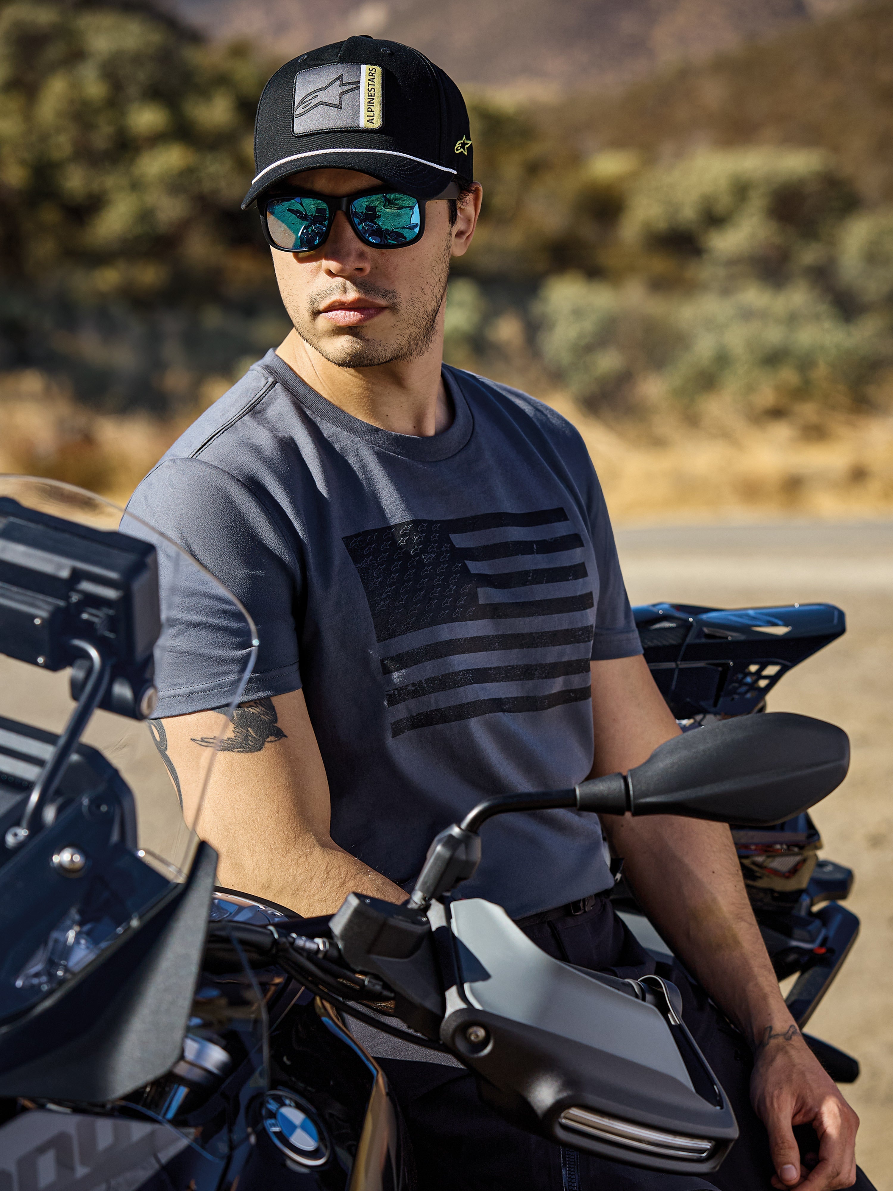 Alpinestars Waving CSF Tee, kortærmet t-shirt, koksgrå, med sort grafik af et bølgende flag på brystet, båret af en mand på en motorcykel i solrige udendørs omgivelser, afslappet motorsport-livsstil