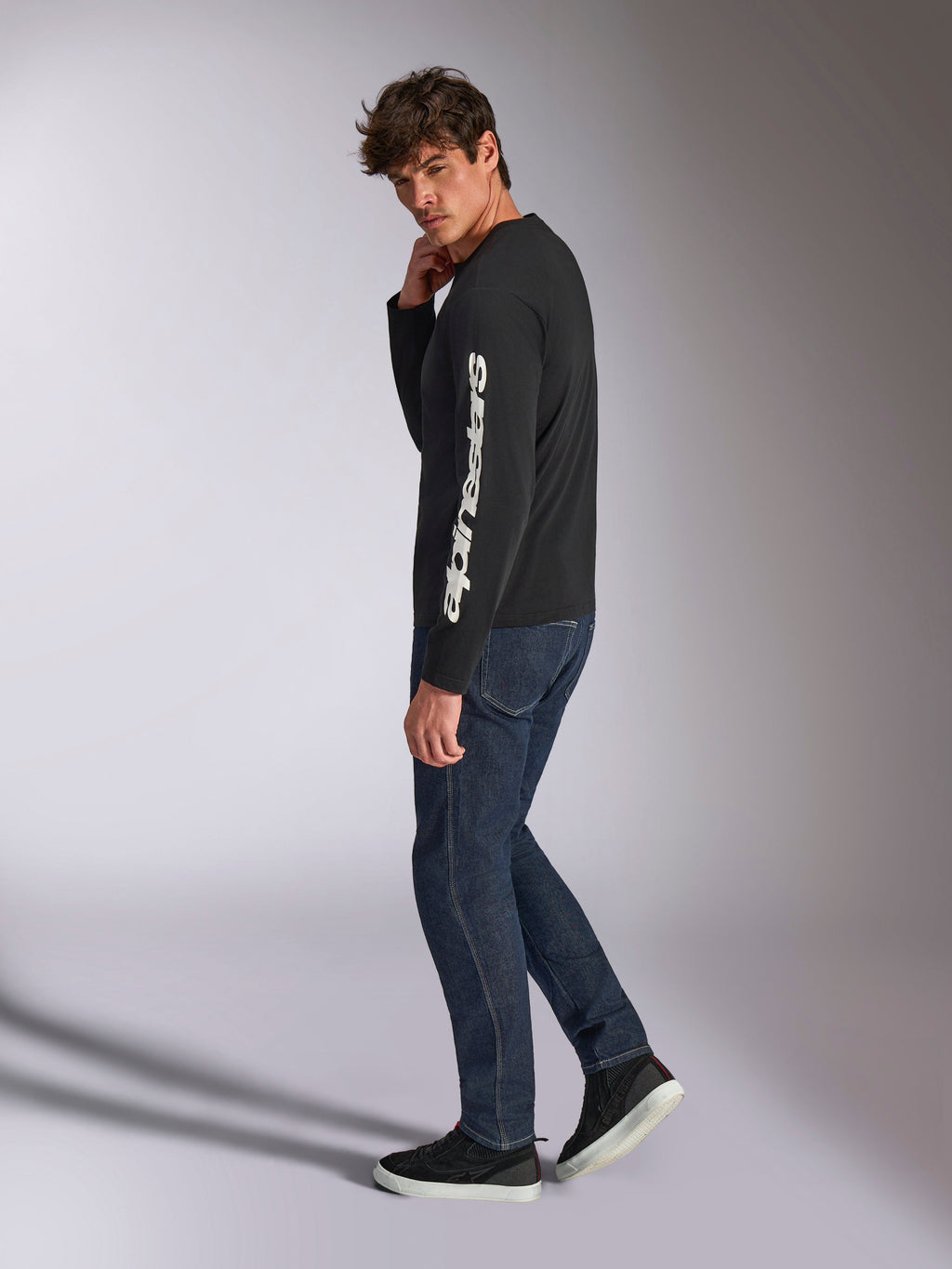Alpinestars Trackside CSF langærmet t-shirt, sort med hvidt lodret Alpinestars-tekstlogo på ærmet, vist på en model med mørke denimjeans og sneakers, afslappet motorsport-inspireret sportstøj