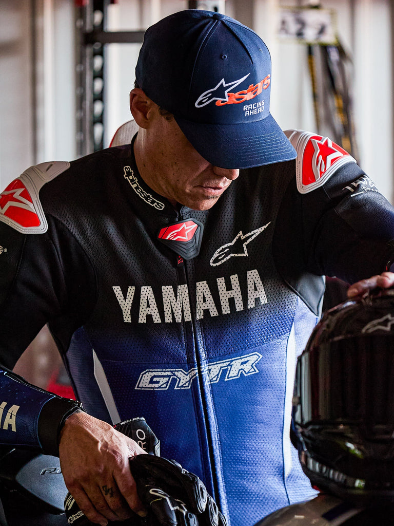 Alpinestars Entitled Hat, Navy Blue with Orange Astars logo and white 'Racing Ahead' text, worn by a rider in a blue and black Yamaha racing suit, curved bill baseball cap with structured crown