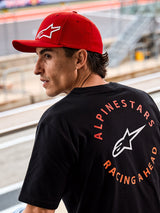 Alpinestars Corp Shift Multi kasket, rød kasket med buet skygge og hvidt 3D Astars-logo, båret sammen med en sort Alpinestars t-shirt med 'Racing Ahead'-grafik, lifestyle motorsport-miljø ved en racerbane