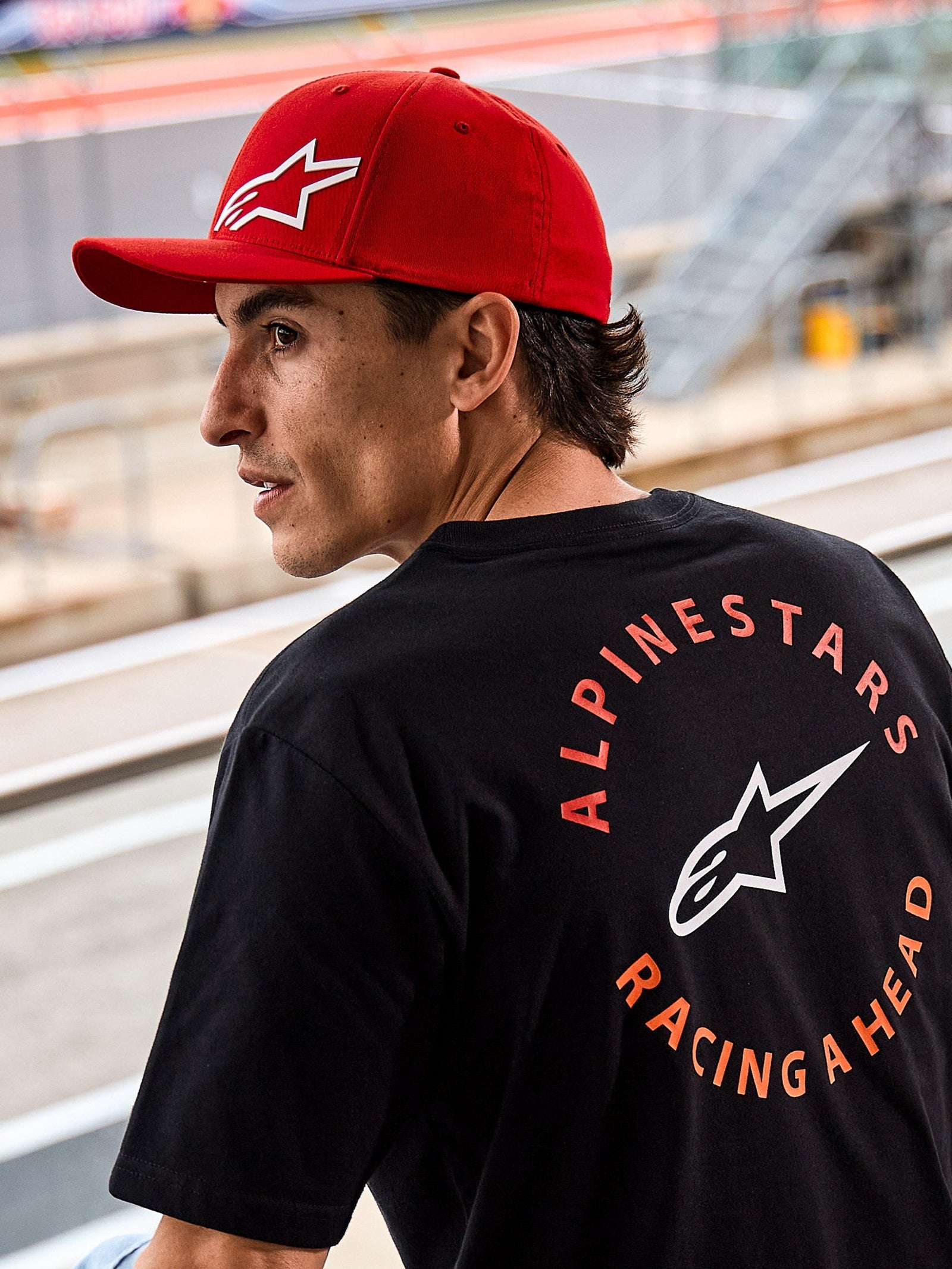Alpinestars Corp Shift Multi kasket, rød kasket med buet skygge og hvidt 3D Astars-logo, båret sammen med en sort Alpinestars t-shirt med 'Racing Ahead'-grafik, lifestyle motorsport-miljø ved en racerbane