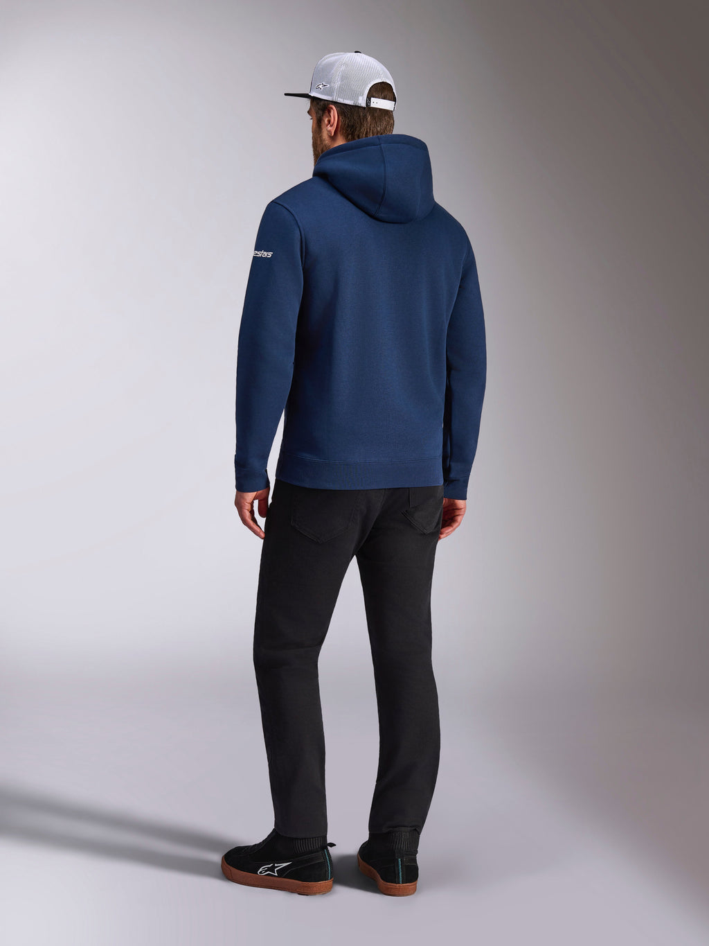 Alpinestars Frontal Hoodie, mørkeblå pullover fleece hættetrøje set bagfra, hvidt tekstlogo på ærmet, sorte bukser og hvid mesh trucker kasket, afslappet motorsport-inspireret streetwear