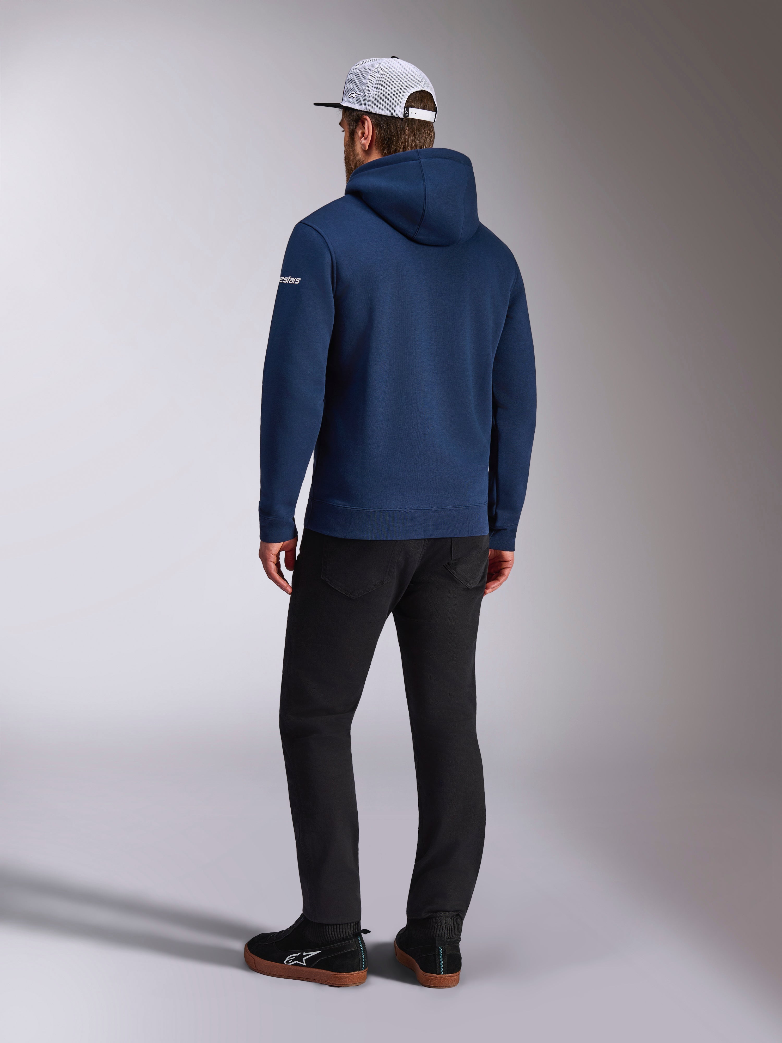 Alpinestars Frontal Hoodie, mørkeblå pullover fleece hættetrøje set bagfra, hvidt tekstlogo på ærmet, sorte bukser og hvid mesh trucker kasket, afslappet motorsport-inspireret streetwear