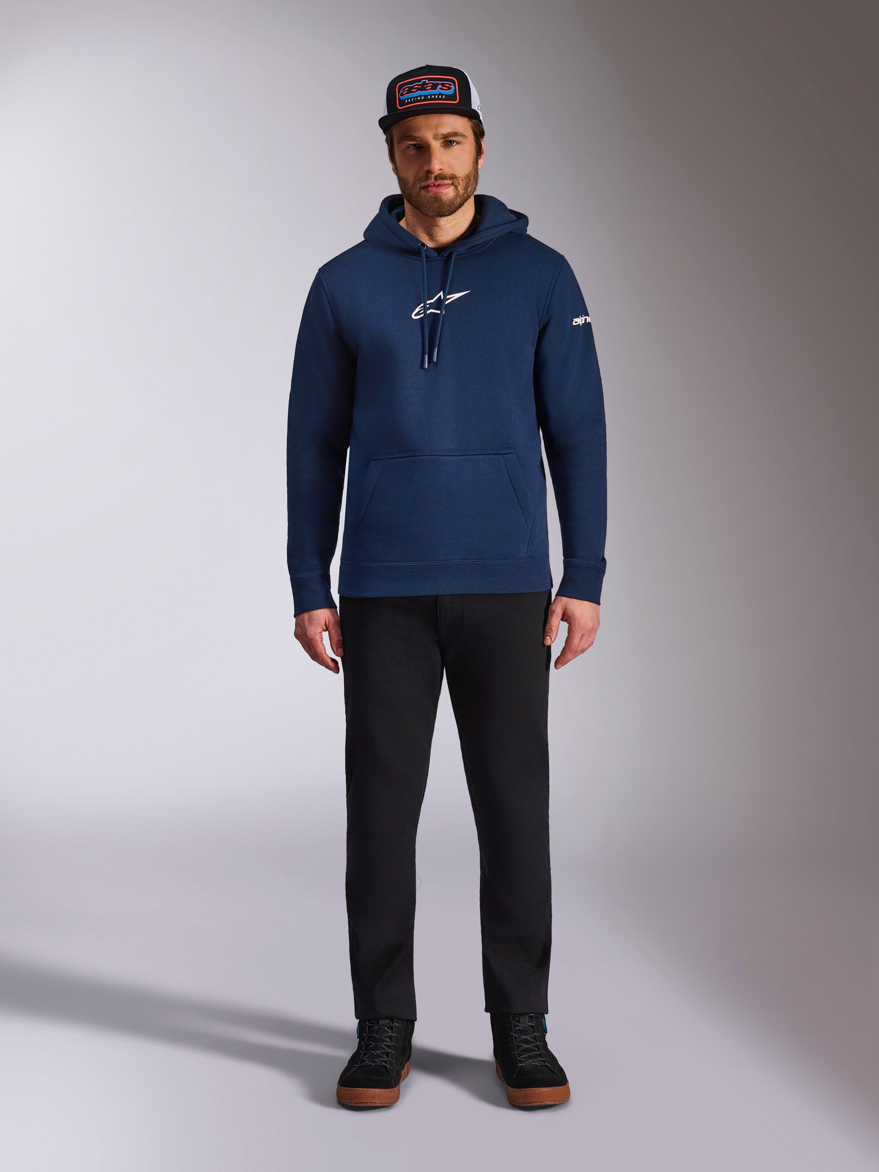 Mandlig model iført Alpinestars Frontal Hoodie i Navy, pullover fleece med hvidt Astars-logo på brystet og tekstlogo på ærmet, kængurulomme og hætte med snøre, kombineret med sorte bukser og kasket med logo, afslappet motorsport-streetwear