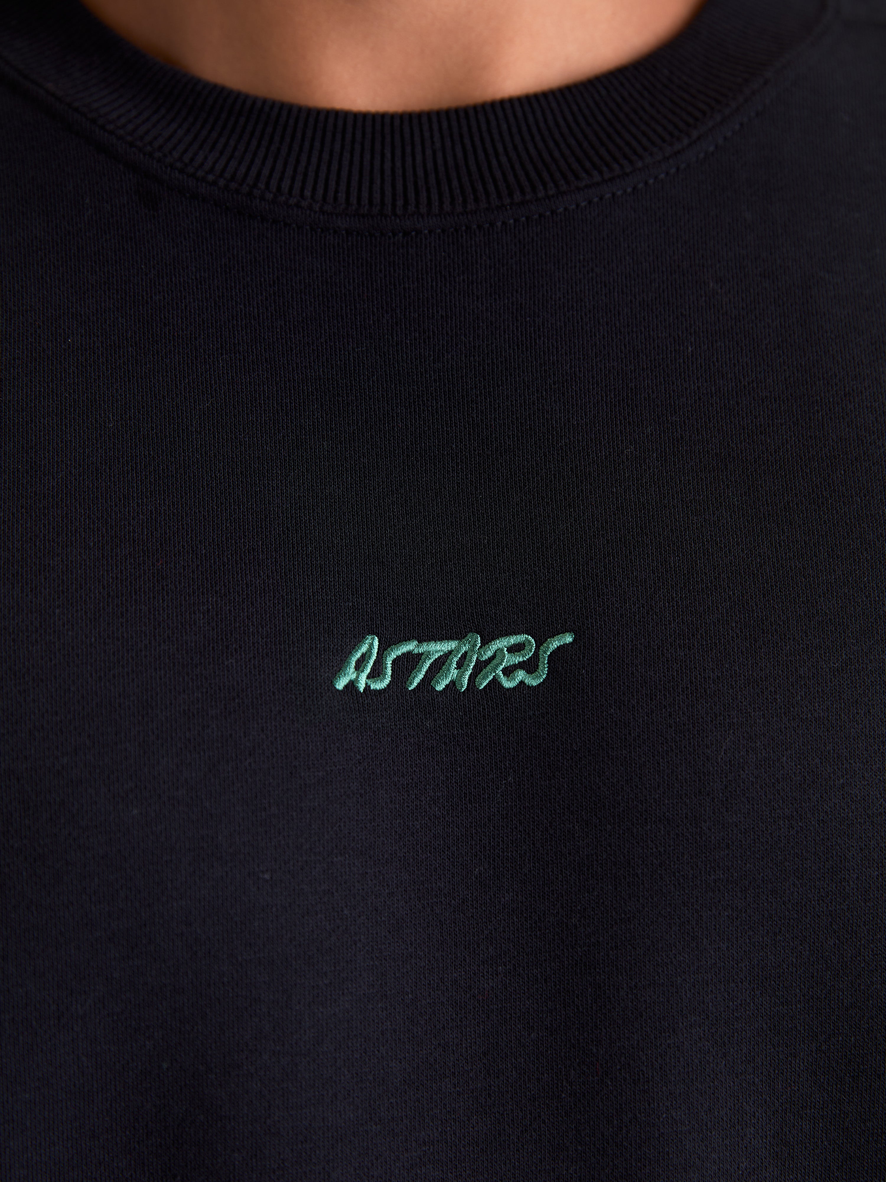 Alpinestars Guise Oversized Crew, sort fleecepullover, nærbillede af den ribstrikkede crew neck og det grønne broderede Astars-logo på brystet, fremstillet i en blød blanding af bomuld og genanvendt polyester.