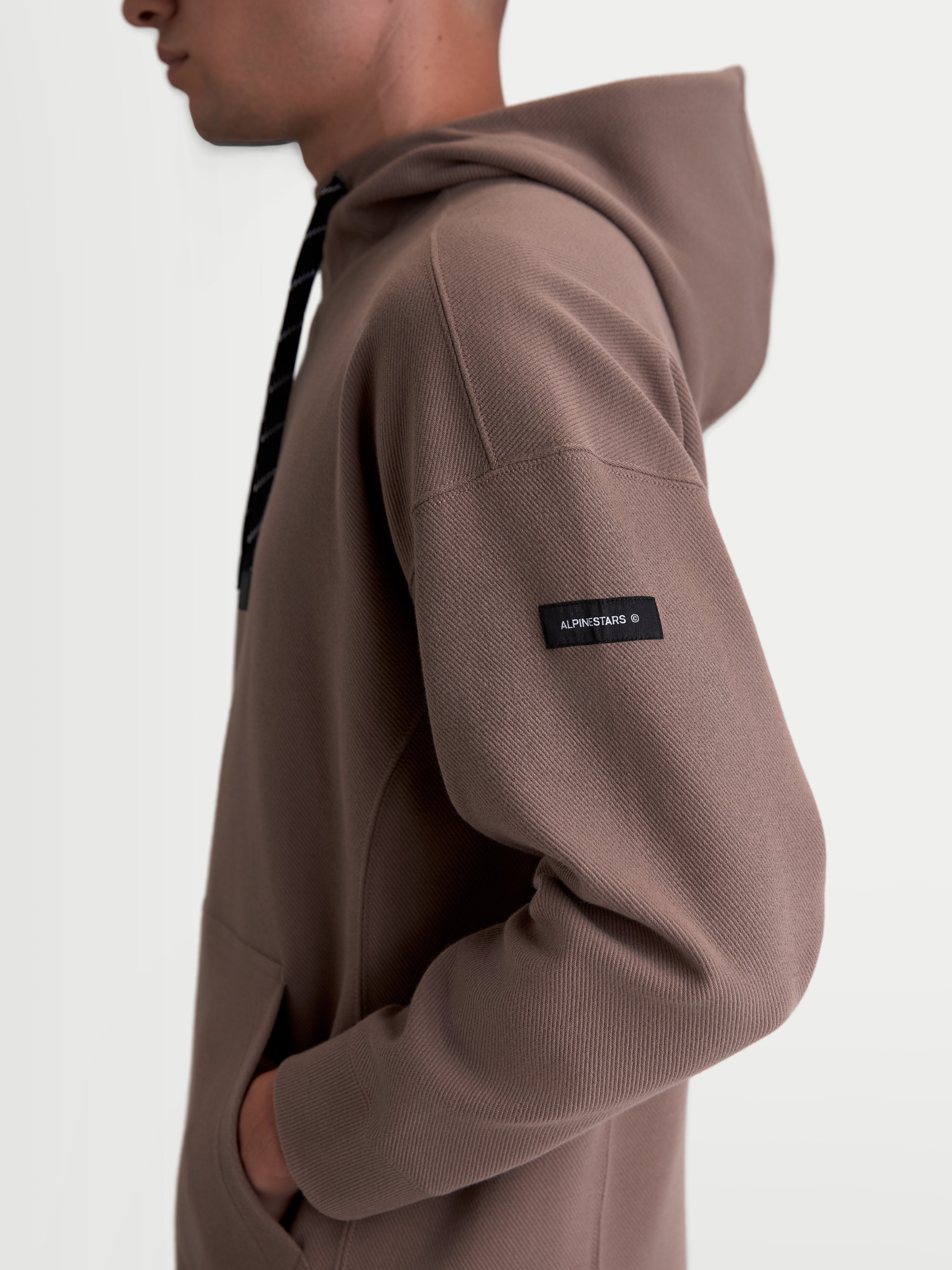 Alpinestars Camino Textured Oversized hættetrøje, brun, tekstureret strikket twill-fleece, sidevisning på model med vævet Alpinestars-label på ærmet, afslappet sportswear.