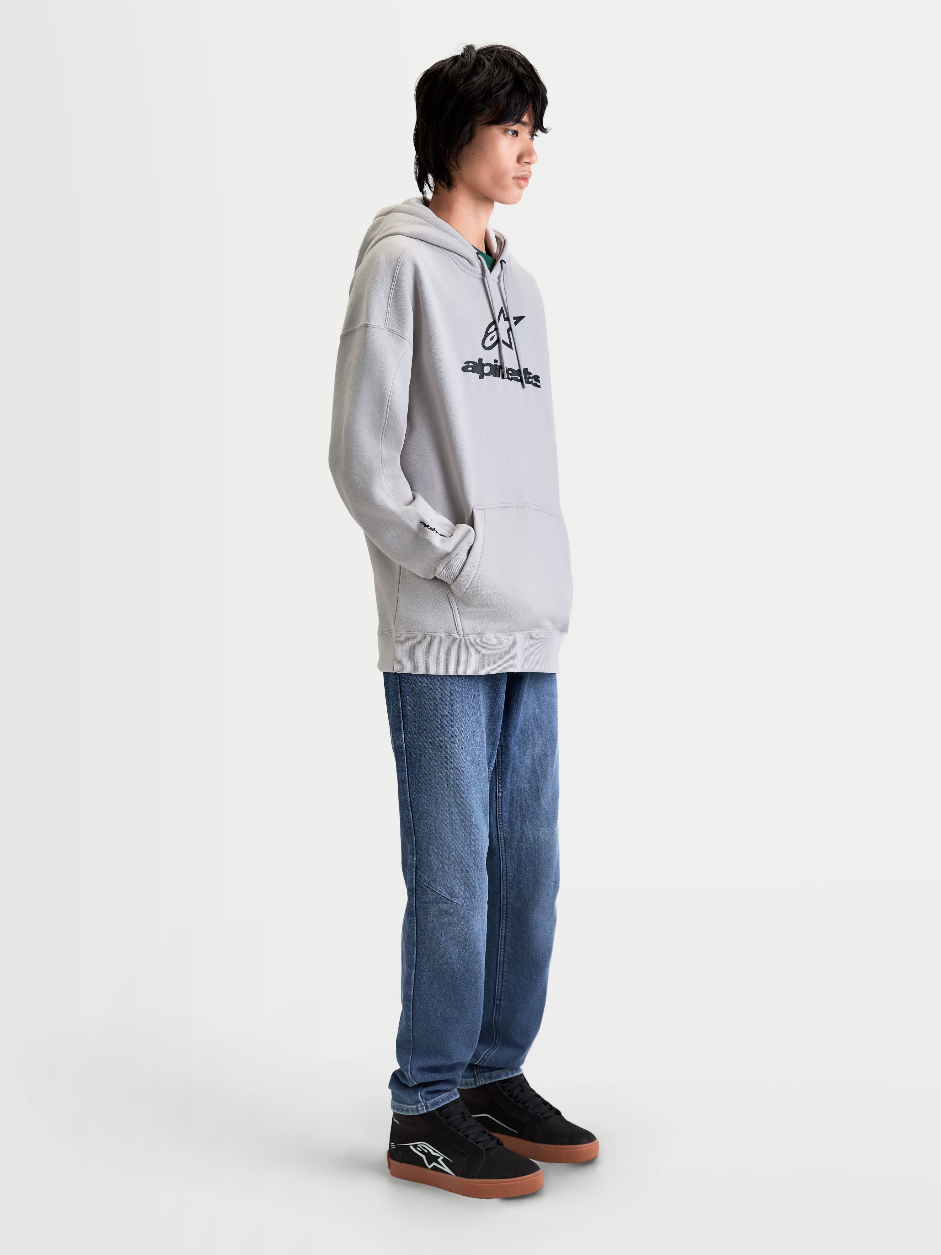 Alpinestars Prelude Oversized Hættetrøje, afslappet herremodel i cementgrå med sort puff-print-logo på brystet, komfortabel fleece-pullover med oversized pasform, kængurulomme og selvforet hætte, vist på model i profil.