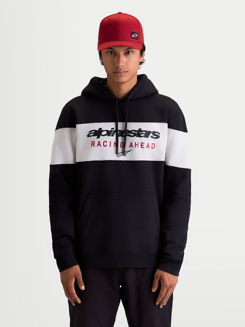 Alpinestars Cubic hættetrøje, herre sportswear pullover fleece i sort og hvidt farveblokeret design, Alpinestars Racing Ahead-logo på brystet, kængurulomme og snøre i hætten, afslappet livsstilstøj set på model med rød kasket.