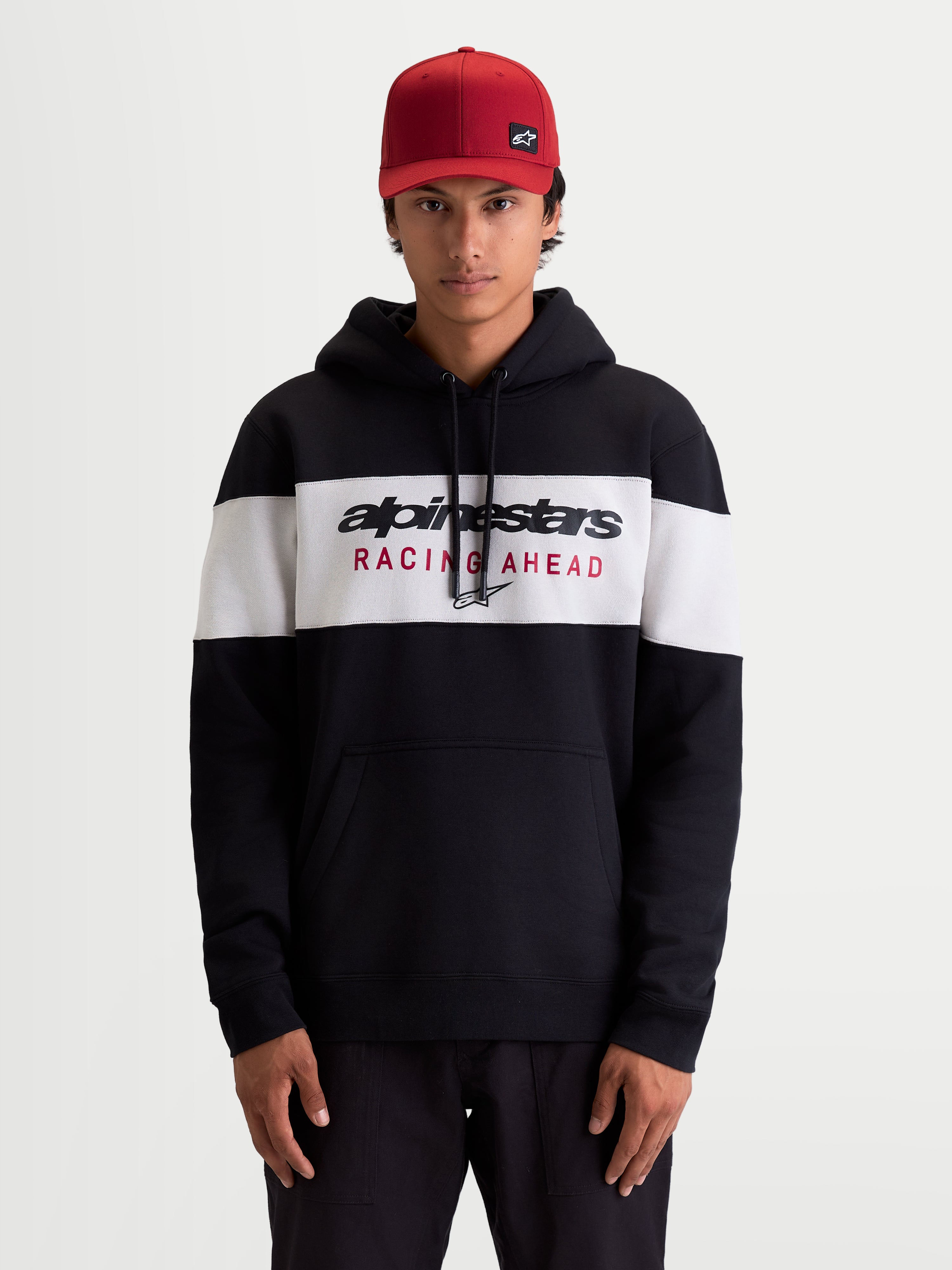 Alpinestars Cubic hættetrøje, herre sportswear pullover fleece i sort og hvidt farveblokeret design, Alpinestars Racing Ahead-logo på brystet, kængurulomme og snøre i hætten, afslappet livsstilstøj set på model med rød kasket.