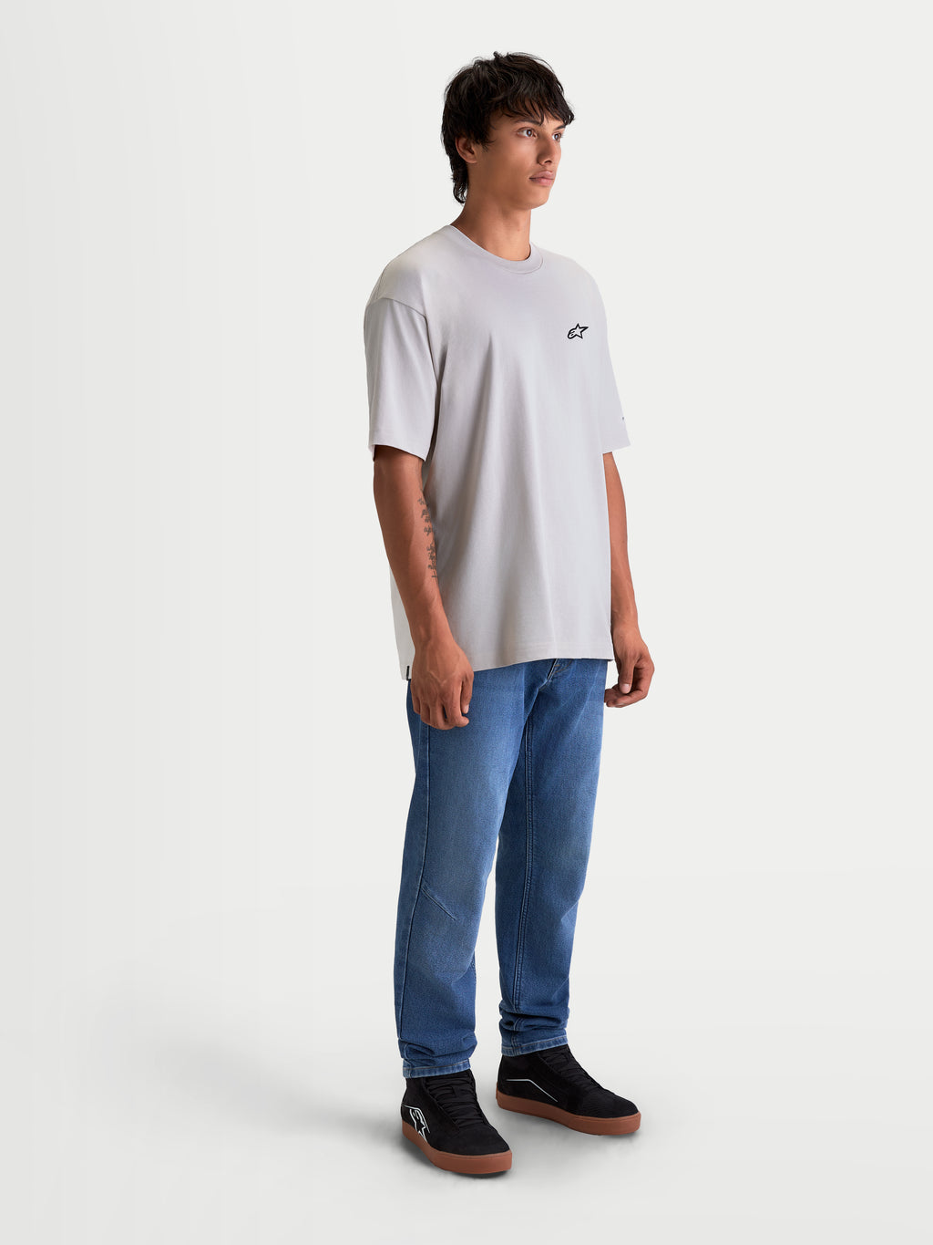 Alpinestars Mandate Oversized CSF T-shirt, oversized kortærmet sportswear t-shirt, cementgrå, med lille sort Astars-logo på brystet. Afslappet pasform i blød bomulds-polyesterblanding til hverdagsbrug. Vist på en mandlig model iført blå jeans og sorte sneakers.