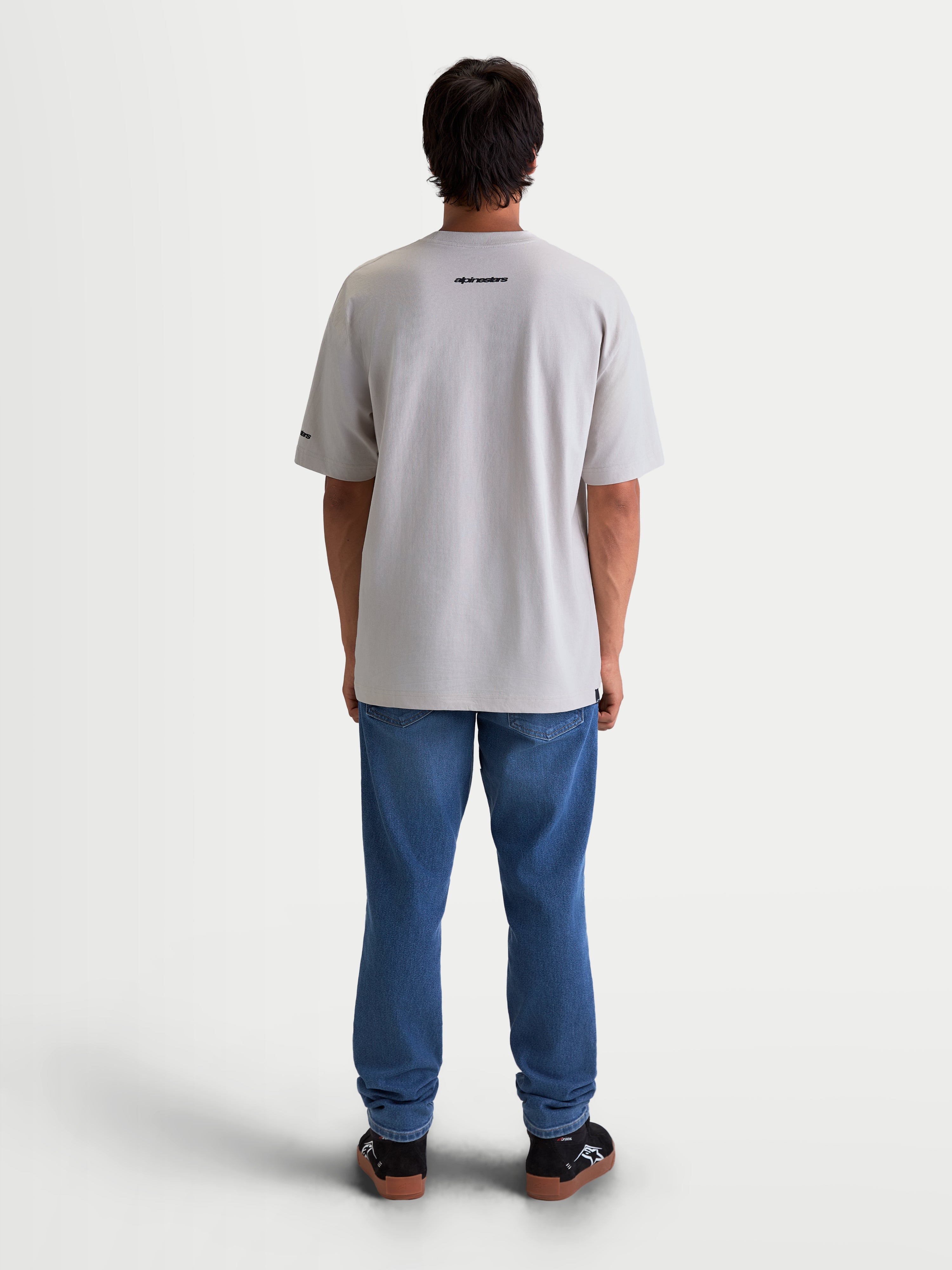 Alpinestars Mandate Oversized CSF T-shirt, unisex sportswear, cementgrå, bagfra på model. Afslappet oversized kortærmet t-shirt med sort Alpinestars-ordgrafik på ryggen, fremstillet i en blød bomulds- og polyesterblanding.