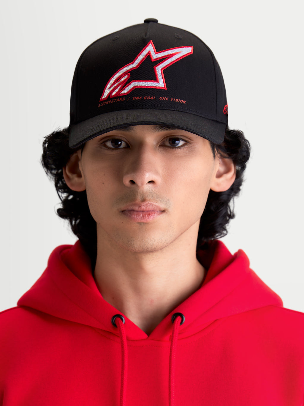 Alpinestars Lineout kasket, sportswear baseballkasket med buet skygge, sort med rødt og hvidt broderet Astars-logo og One Goal One Vision-branding, set forfra på model iført en rød hættetrøje, struktureret twill-konstruktion.