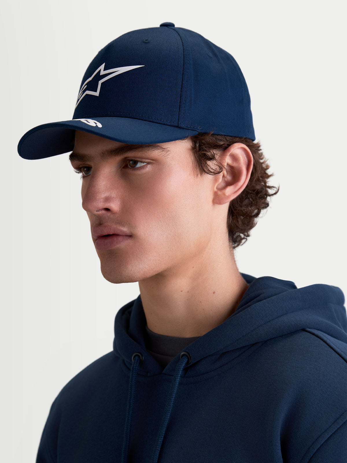 Alpinestars Arclight kasket, navyblå sportswearkasket til mænd og kvinder, hvidt Astars puff-print logo, buet skygge og struktureret krone i bomuldstwill, sideprofil på model med et klassisk motorsportsinspireret look.