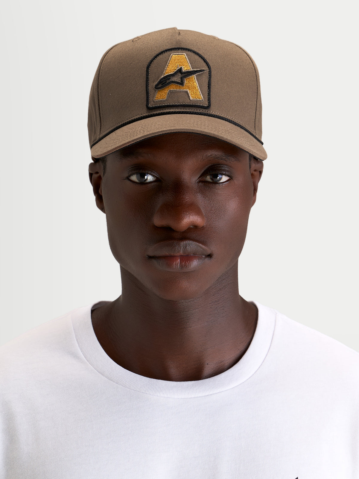 Alpinestars Acme Snapback Kasket, brun sportswear-kasket, båret af en model, med struktureret krone, buet skygge og broderet Astars-logopatch med rebkant foran