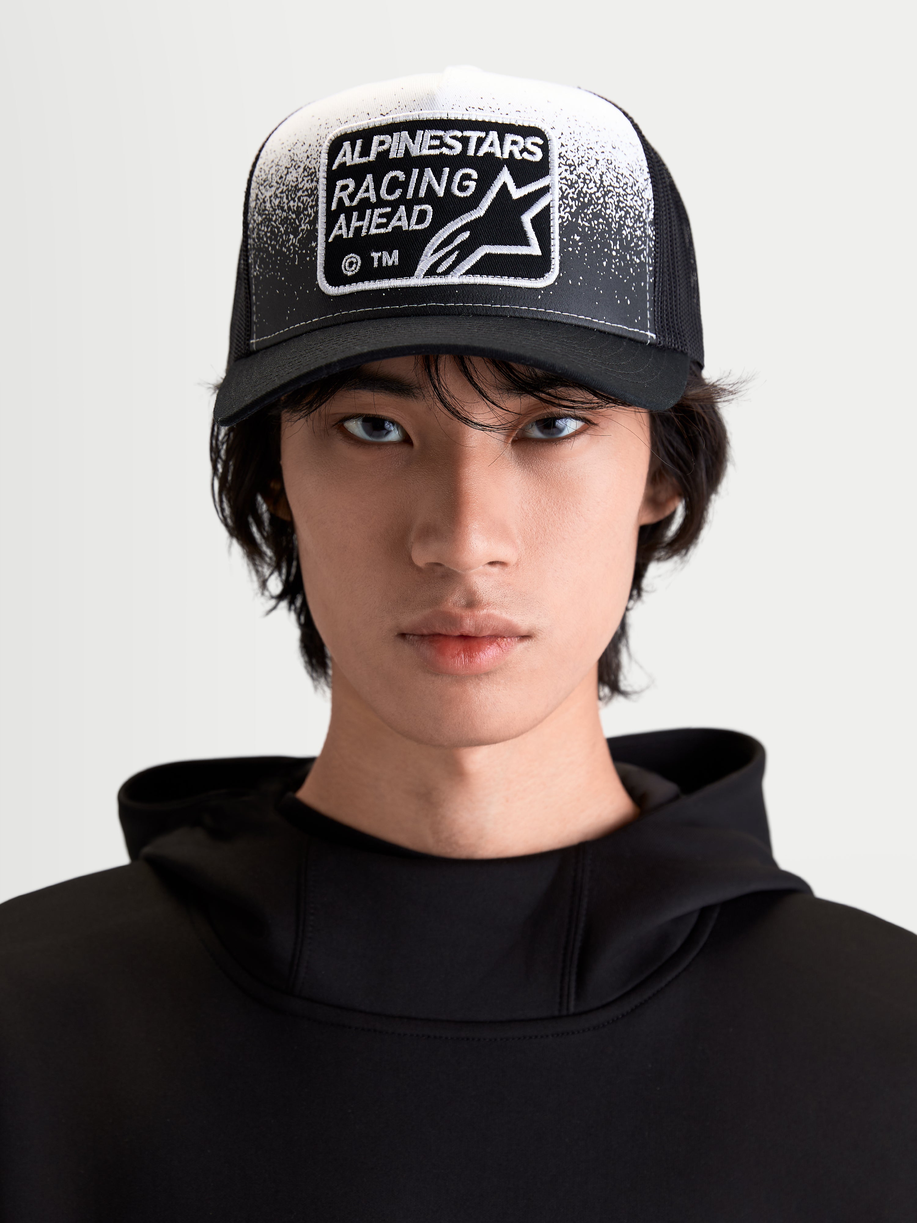 Alpinestars Gravel Trucker cap, livsstilskasket til sportstøj, sort og hvid, set forfra på model med buet skygge, åndbar mesh-bagside og vævet Racing Ahead-patch med grusstruktur-detaljer.