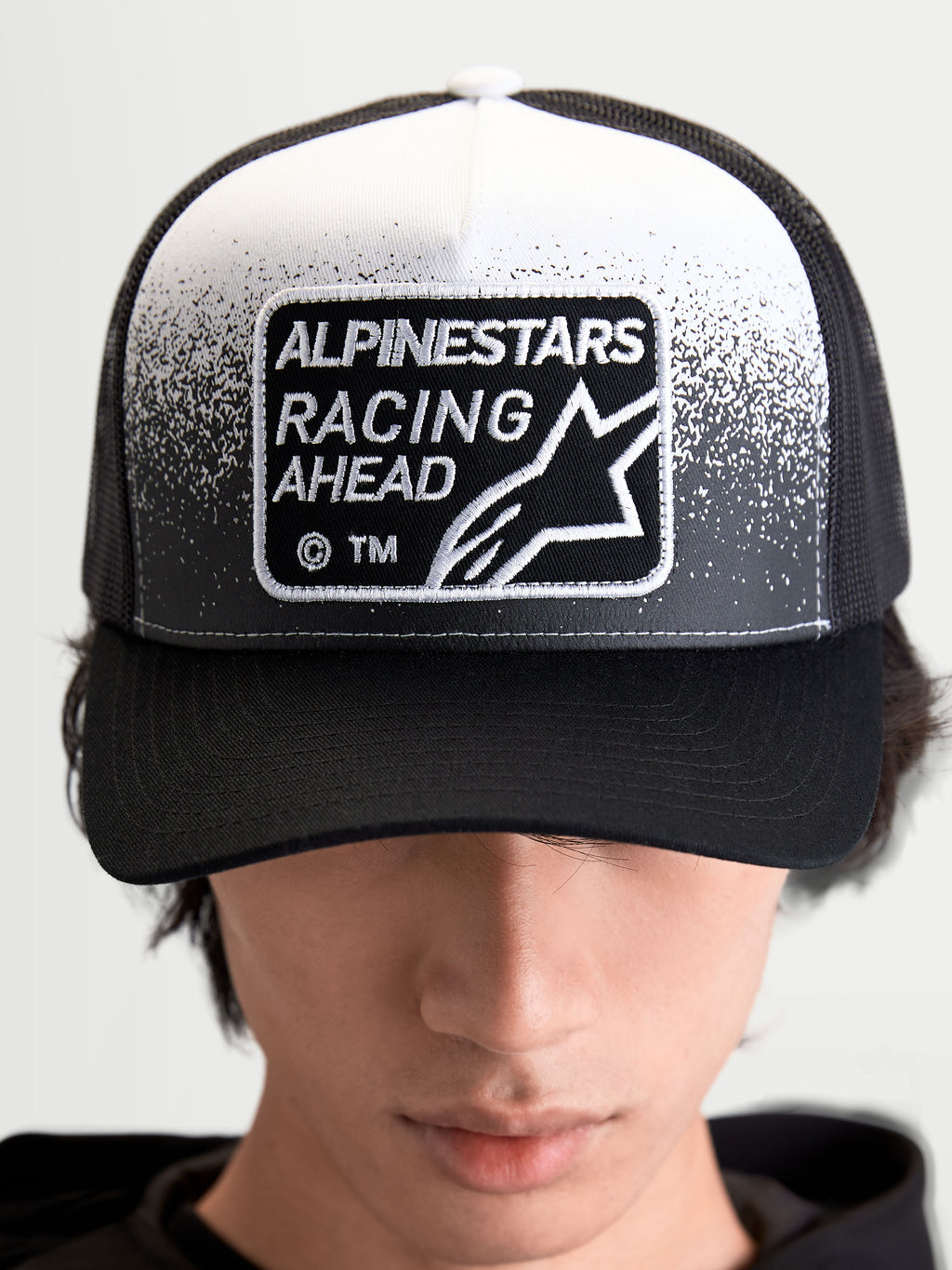 Alpinestars Gravel Trucker kasket, sportswear trucker cap i sort og hvid med et vævet Alpinestars Racing Ahead-mærke med grusstruktur-detaljer, struktureret krone og buet skygge, set forfra på model.