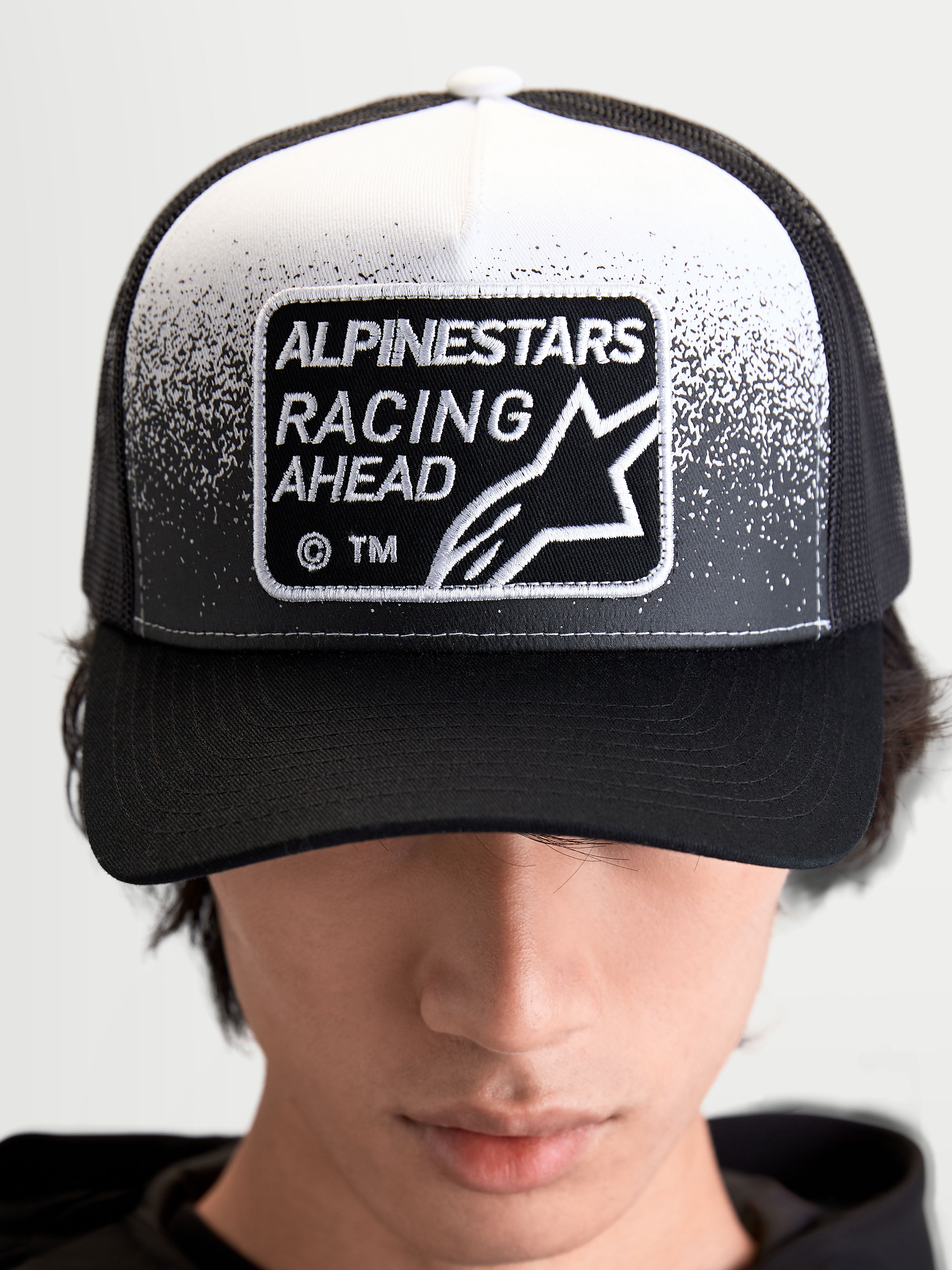 Alpinestars Gravel Trucker kasket, sportswear trucker cap i sort og hvid med et vævet Alpinestars Racing Ahead-mærke med grusstruktur-detaljer, struktureret krone og buet skygge, set forfra på model.