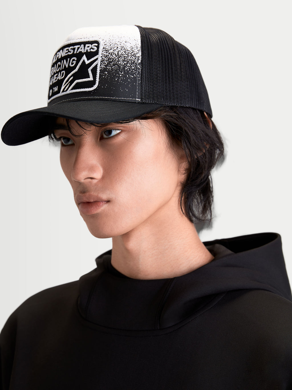 Alpinestars Gravel Trucker cap, afslappet kasket til sportstøj på model, sort og hvid, med struktureret krone, buet skygge og åndbar mesh-bagside. Forsiden har et vævet patch med grusstruktur-grafik og Alpinestars Racing Ahead-branding.