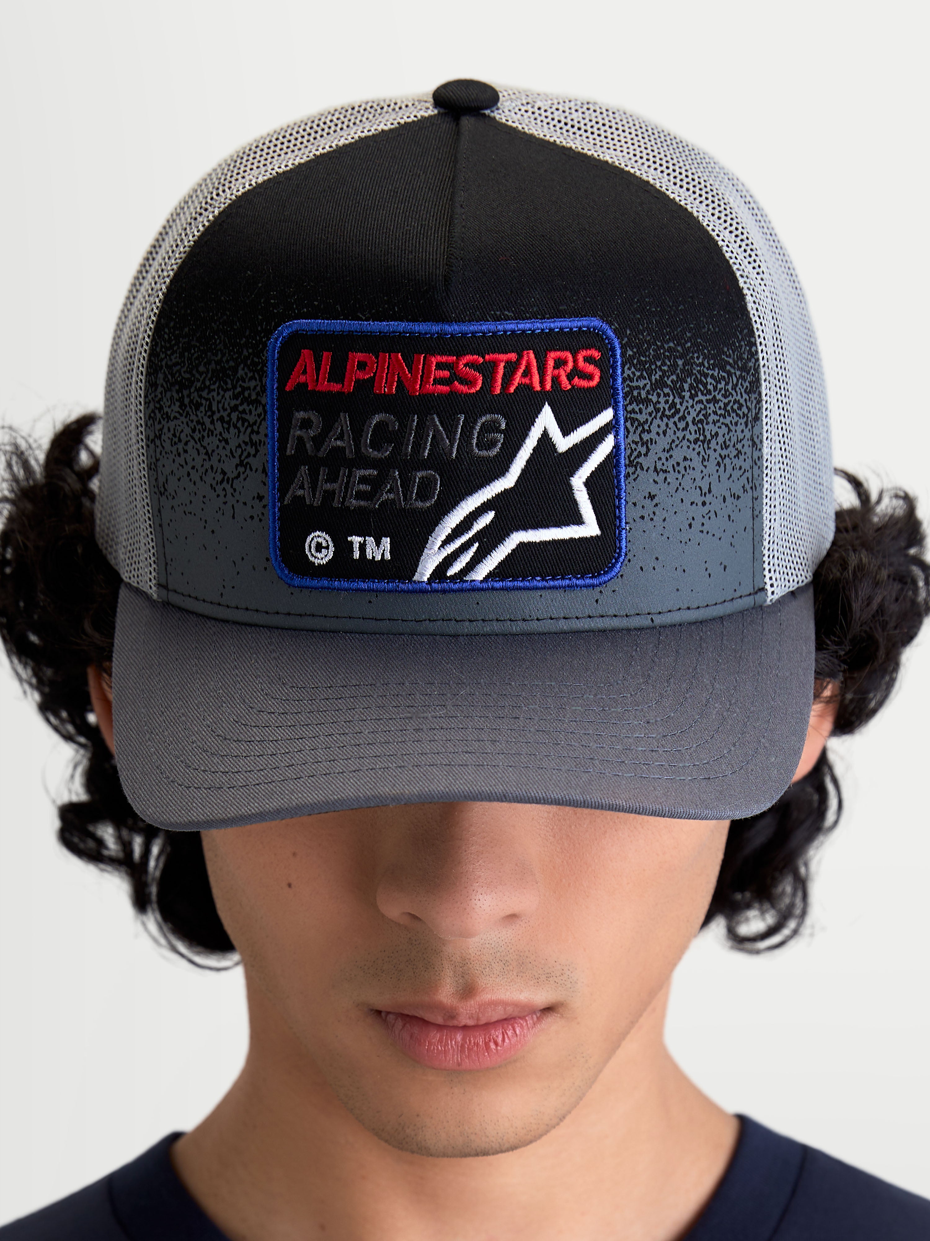 Alpinestars Gravel Trucker Cap, mørkegrå og sort truckerkasket, buet skygge og vævet frontpatch med 'Racing Ahead'-tekst og Astars-logo, vist på model set forfra.