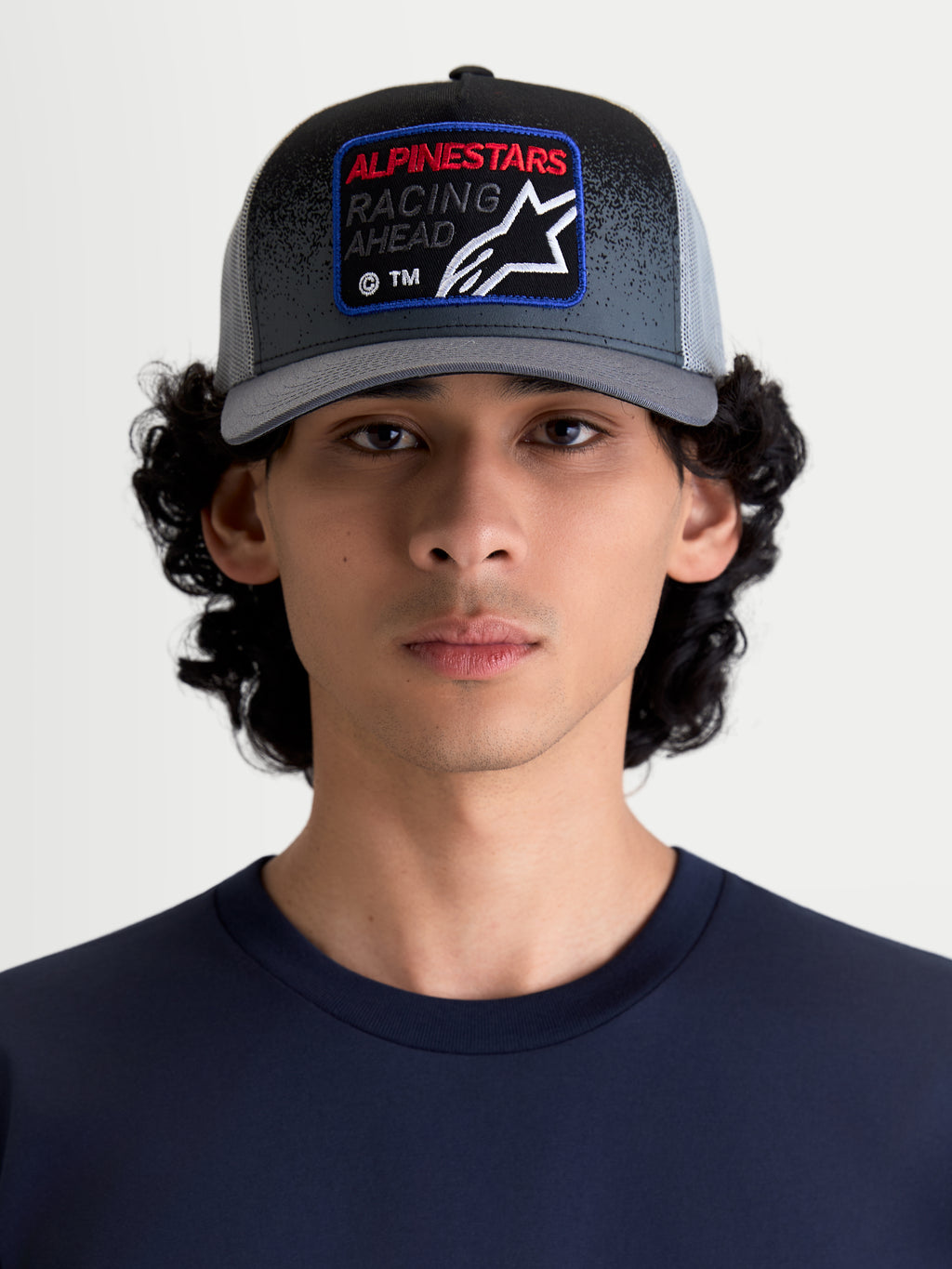 Alpinestars Gravel Trucker Cap, casual trucker kasket til herrer og damer, mørkegrå og sort, vist på en model. Kasketten har en struktureret krone, buet skygge og et vævet frontmærke med Alpinestars Racing Ahead-logo og detaljer med grusstruktur.