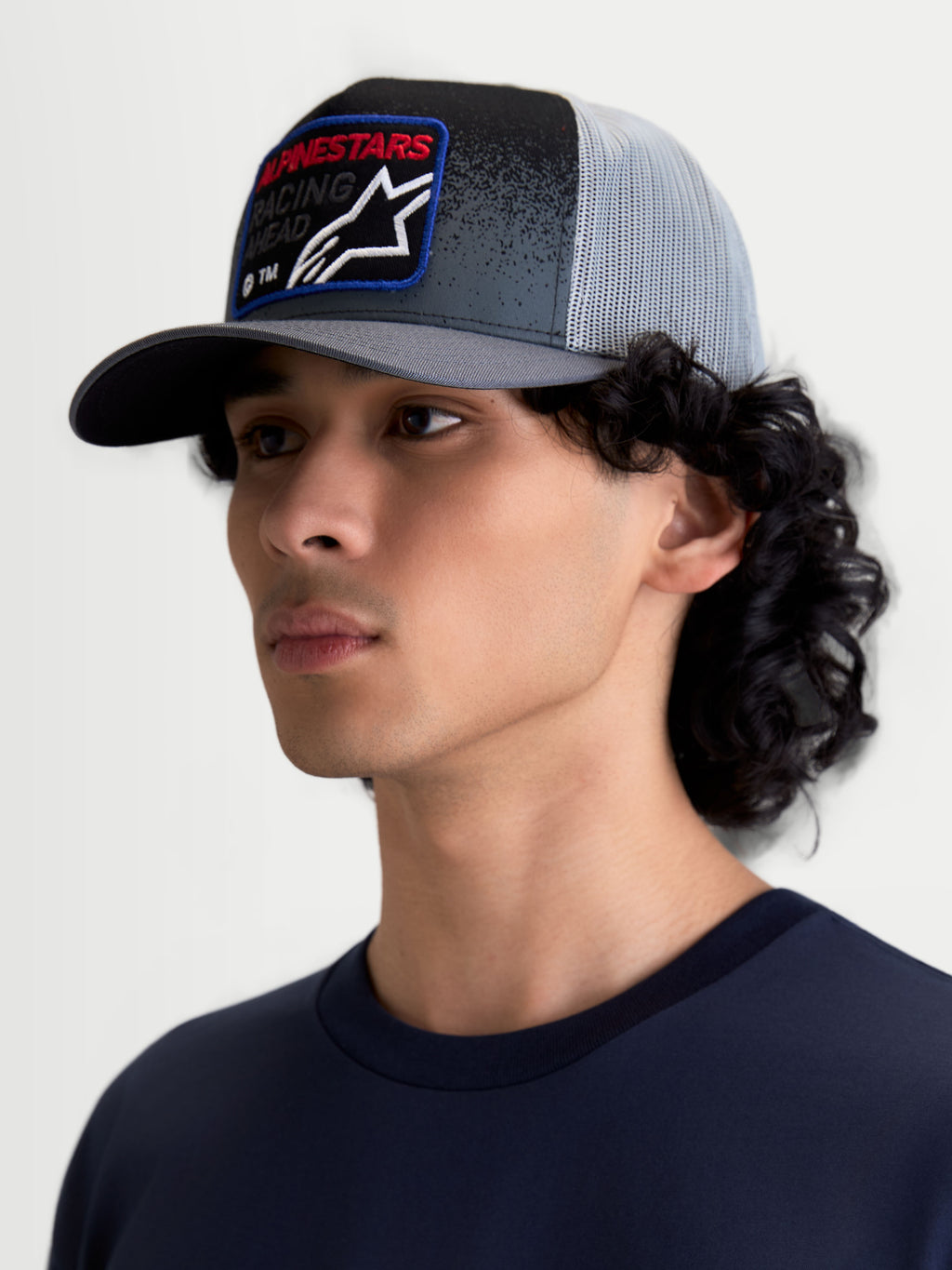 Alpinestars Gravel Trucker cap, mørkegrå og sort kasket vist på model med lysegrå mesh-bagside, struktureret krone, buet skygge og vævet frontpatch med rødt Alpinestars-logo, motorsport-inspireret sportstøj til hverdagsbrug.