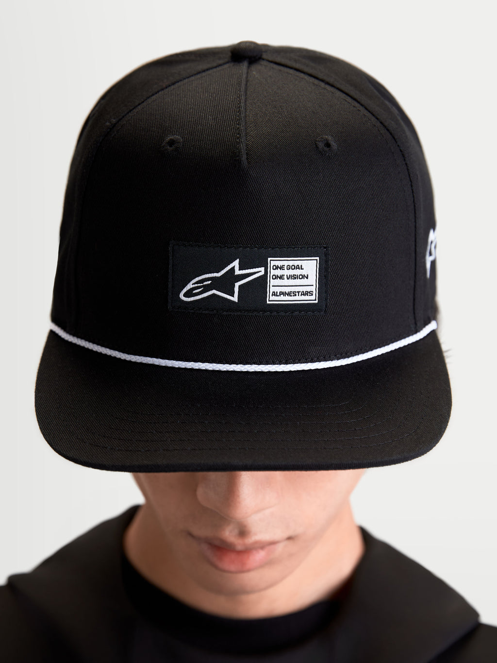Alpinestars Label Snapback kasket, sportswear-kasket til hverdagsbrug, unisex, sort og hvid, bomuldstwill, med struktureret krone, flad skygge, hvid snøre og et vævet mærke foran med Alpinestars branding, vist på model.