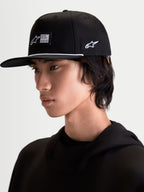 Alpinestars Label Snapback Kasket, sort sportswear-kasket med flad skygge og struktureret krone, vist på model. Denne motorsport-inspirerede kasket har et vævet mærke foran, hvid pyntesnor ved skyggen og broderet logo på siden for et moderne livsstilslook.