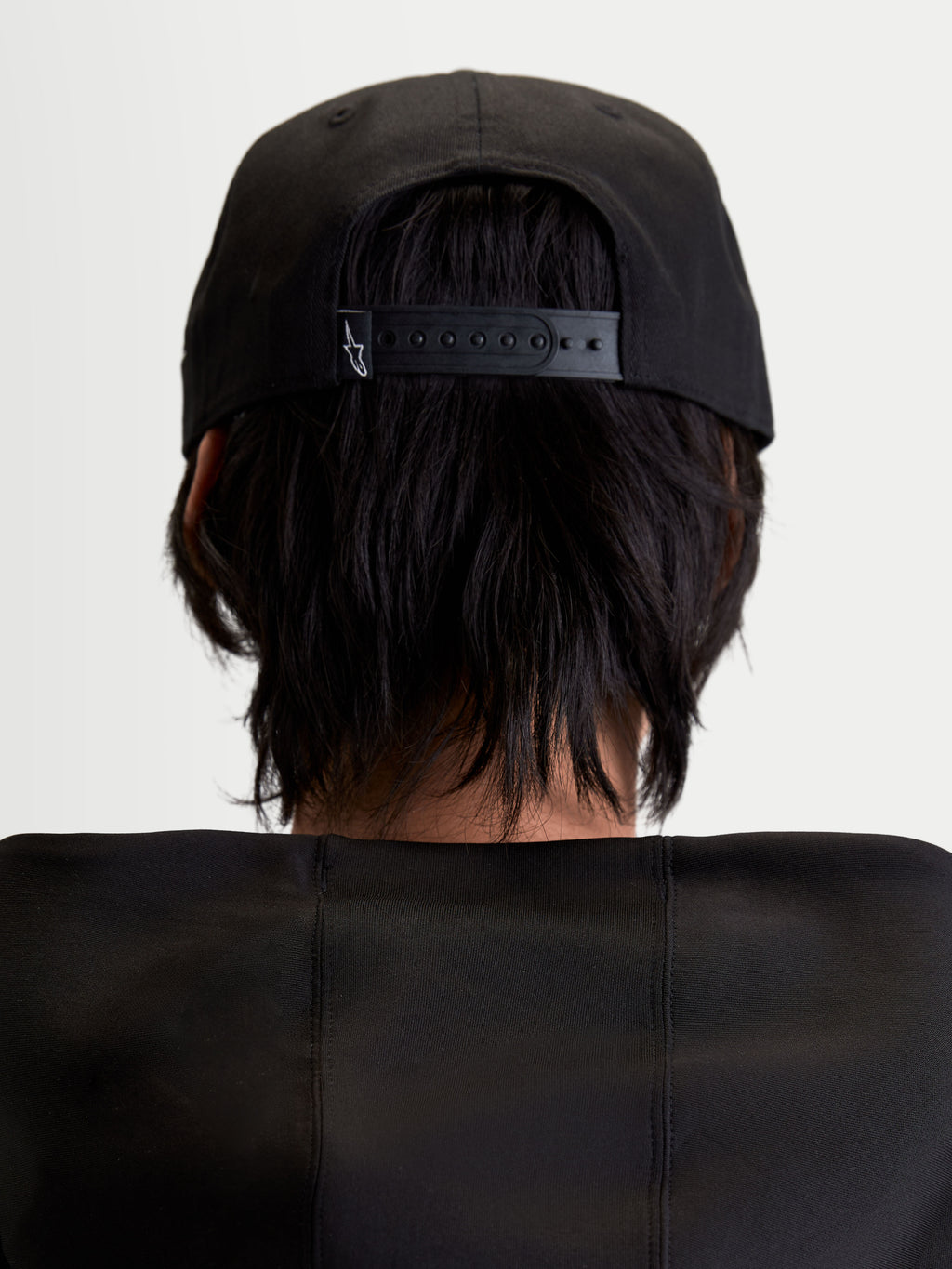 Alpinestars Label Snapback kasket, sort motorsportskasket set bagfra på model, viser den justerbare snapback-lukning med et lille hvidt logo, struktureret bomuldstwill-konstruktion, unisex sportswear.