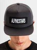 Alpinestars Ten Trucker Cap, mørkegrå sportswear-kasket med flad skygge, båret af model, med et Alpinestars-ordlogo-patch foran og struktureret mesh-krone