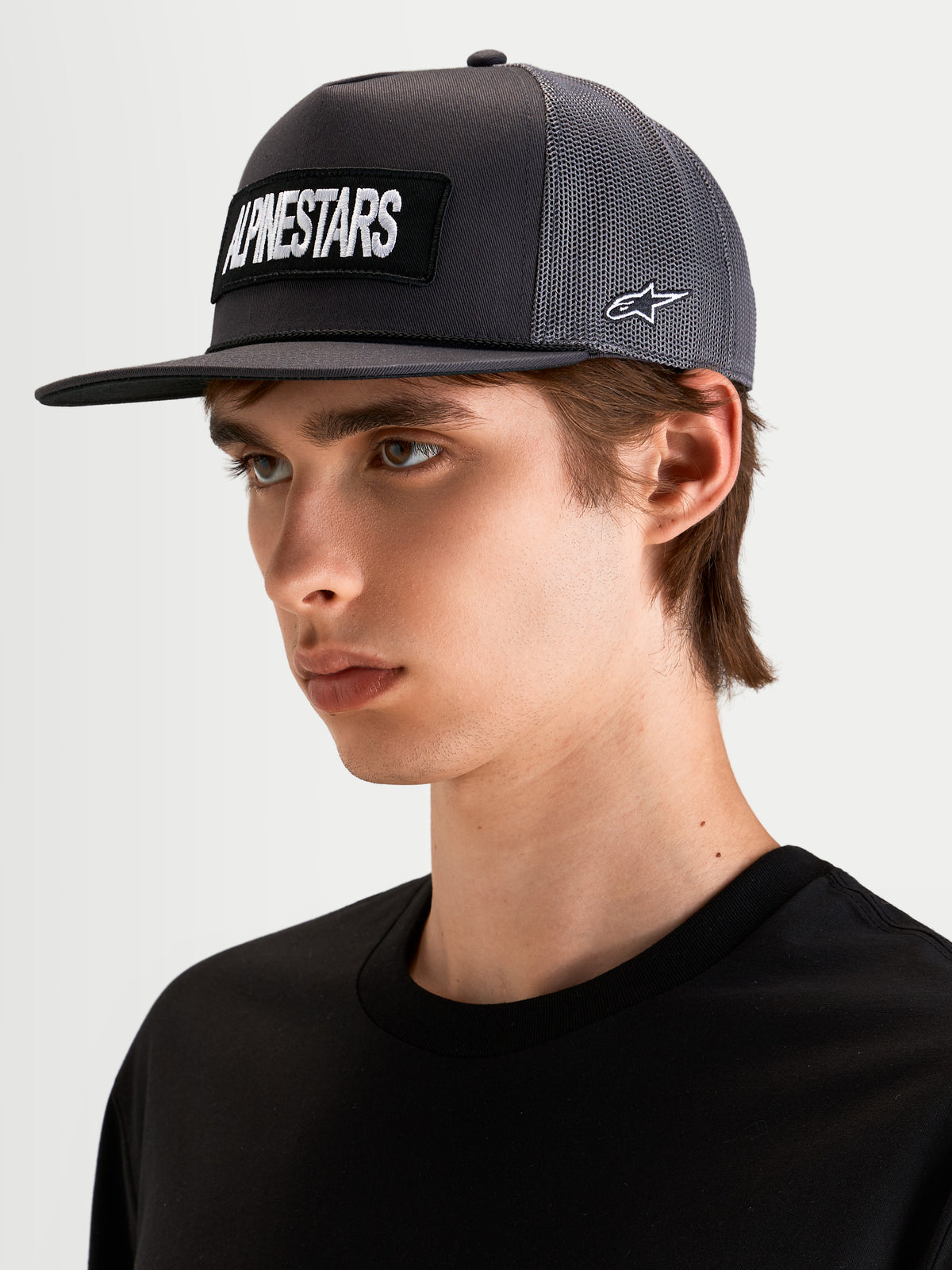 Alpinestars Ten Trucker Cap, mørkegrå sportswear-kasket med flad skygge, båret af model, med broderet Alpinestars-ordlogo-patch foran og Astars-logo i siden
