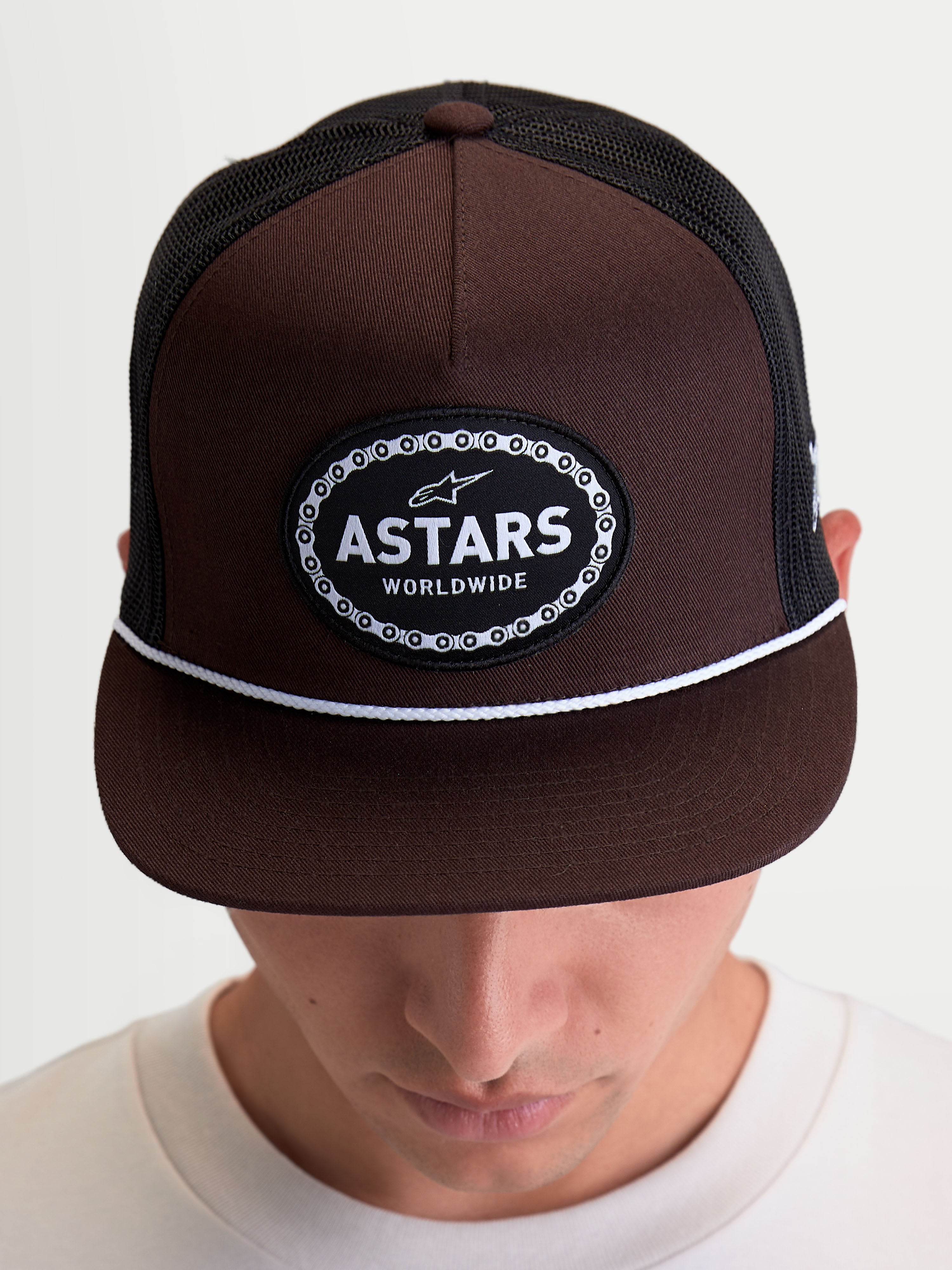 Alpinestars Hotlink Trucker cap, motorsport-inspireret sportswear kasket vist på model, brun og sort, med struktureret krone, flad skygge med hvid snoredetalje og et Astars Worldwide-logomærke med cykelkædekant.
