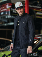 Alpinestars Precedent Jacket, black quilted jacket with Formula 1 styling, featuring an iconic angled quilt pattern, chest logo patch, and white sleeve embroidery, worn with a matching Alpinestars cap in a motorsport garage setting.